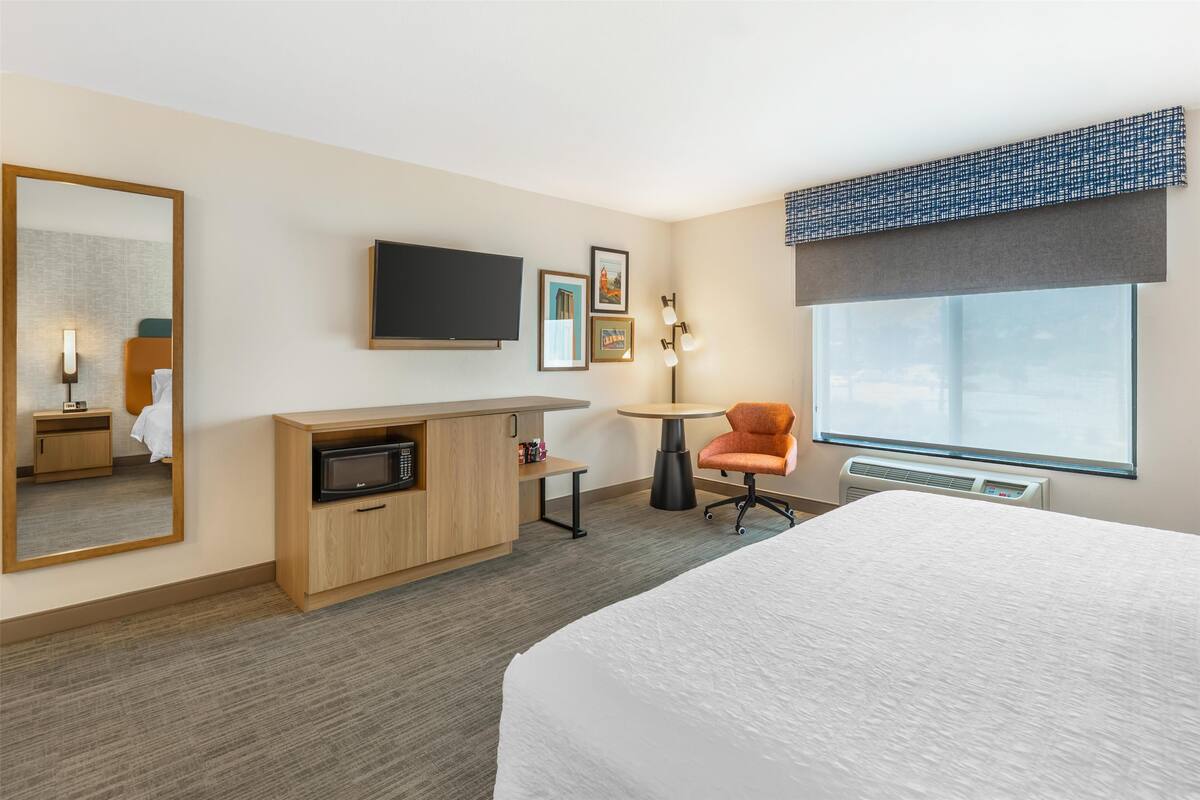 A hotel room with a bed, a desk with a computer, a chair, a TV, and a window with a view.