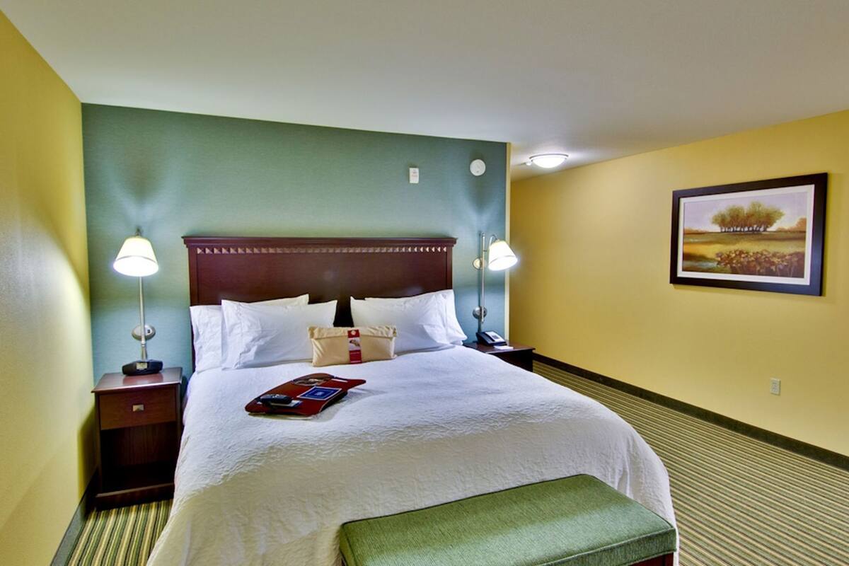 A hotel room with a bed, two bedside tables, a nightstand, a green bench, and a framed picture on the wall.