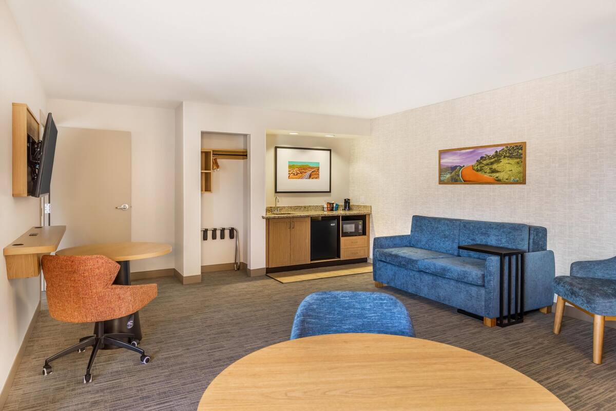 A modern hotel room with a blue sofa, a round wooden table, a small kitchen area, and a TV mounted on the wall.