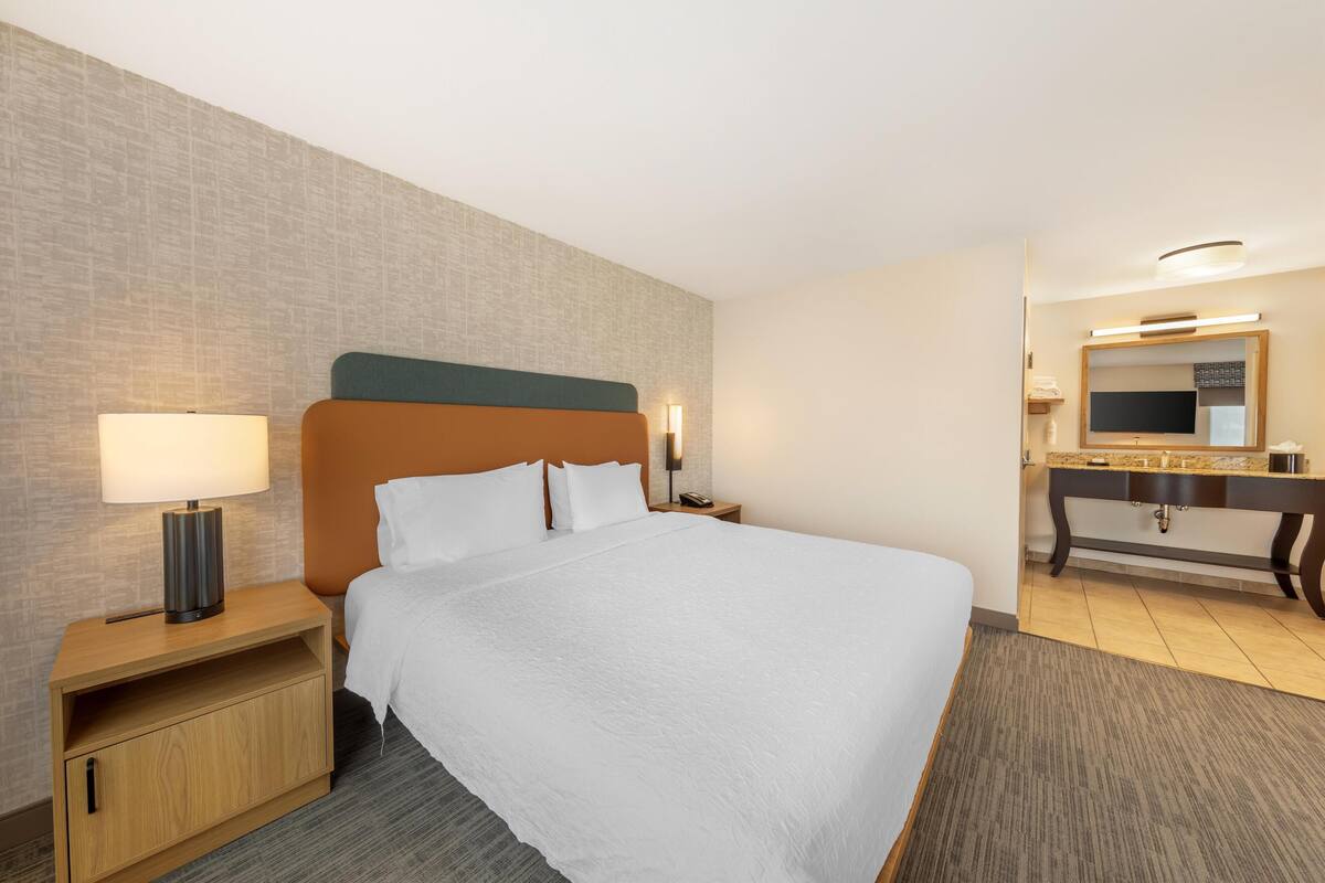 A hotel room with a large bed, a nightstand with a lamp, a wooden dresser, and a mirror above the dresser.