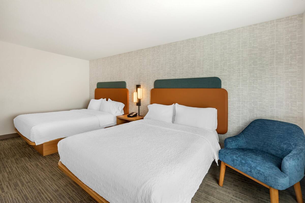 A hotel room with two single beds, a blue armchair, and a nightstand with a lamp.