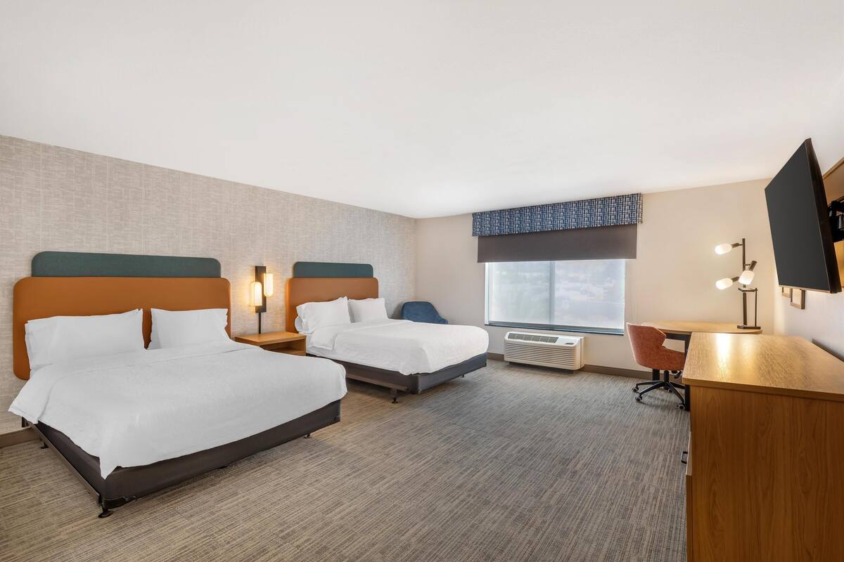 A hotel room with two beds, a desk, a chair, a TV, and a window.