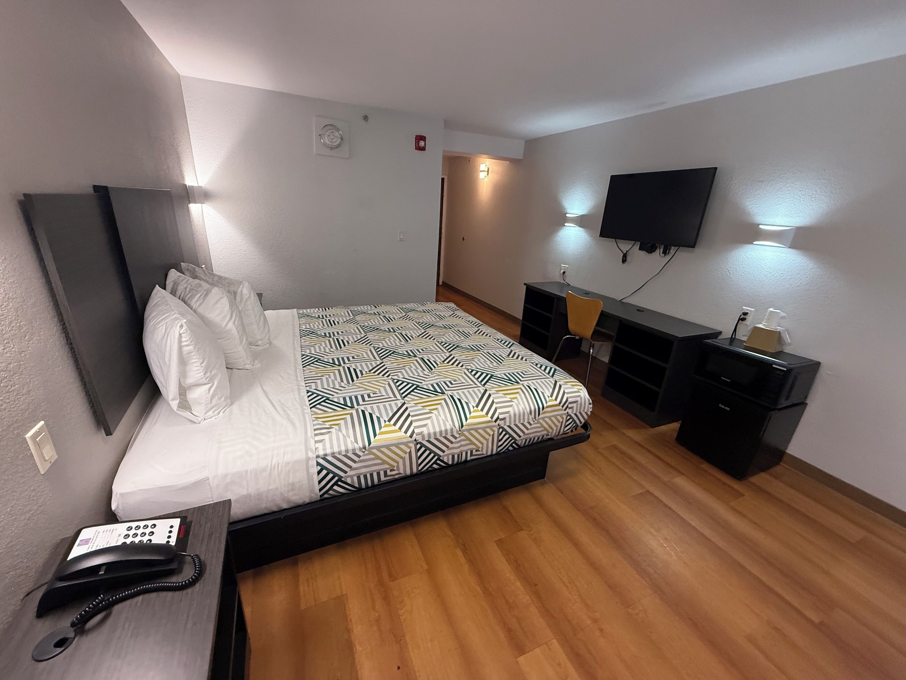 Habitación de hotel con cama, mesita de noche, escritorio con silla, televisor montado en la pared y pequeña cocina.
