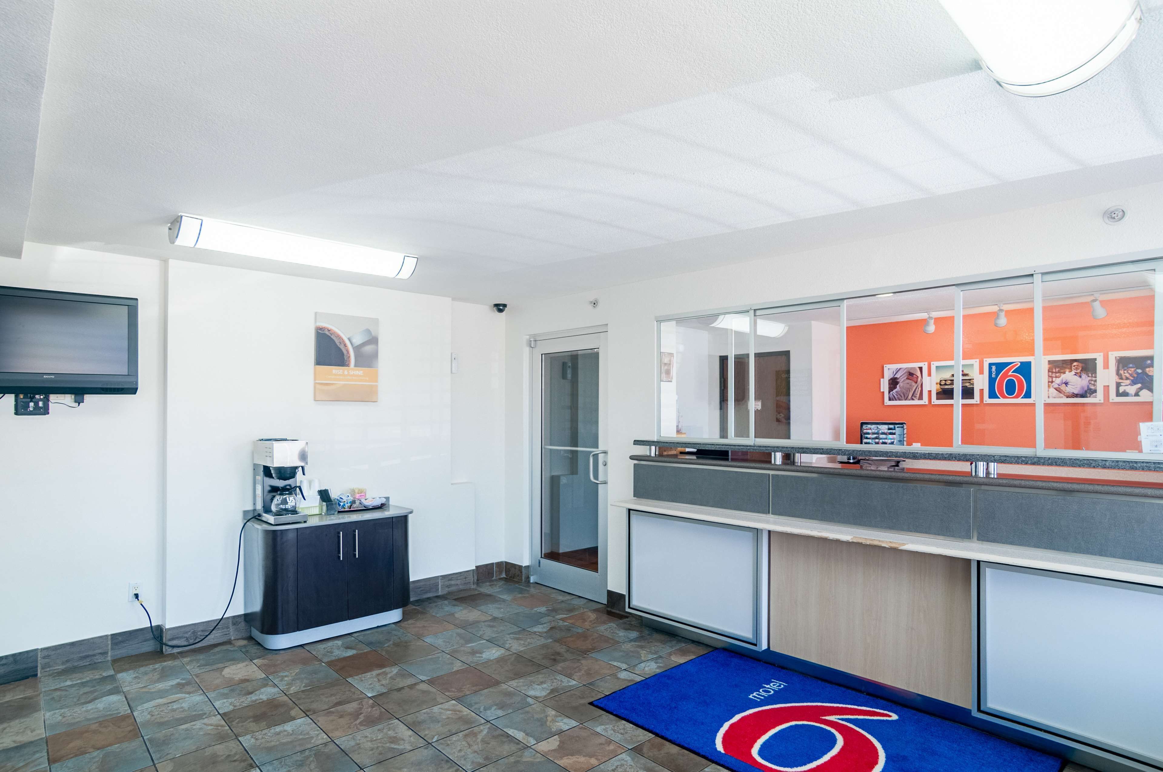 Una recepción moderna con una estación de café, una puerta de cristal que da a una sala, y un mostrador con una decoración en rojo y blanco.