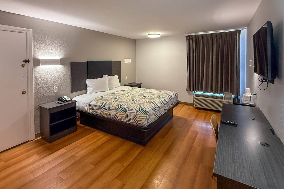Ein Hotelzimmer mit Bett, Nachttisch, Lampe, Klimaanlage und einem an der Wand befestigten Fernseher.