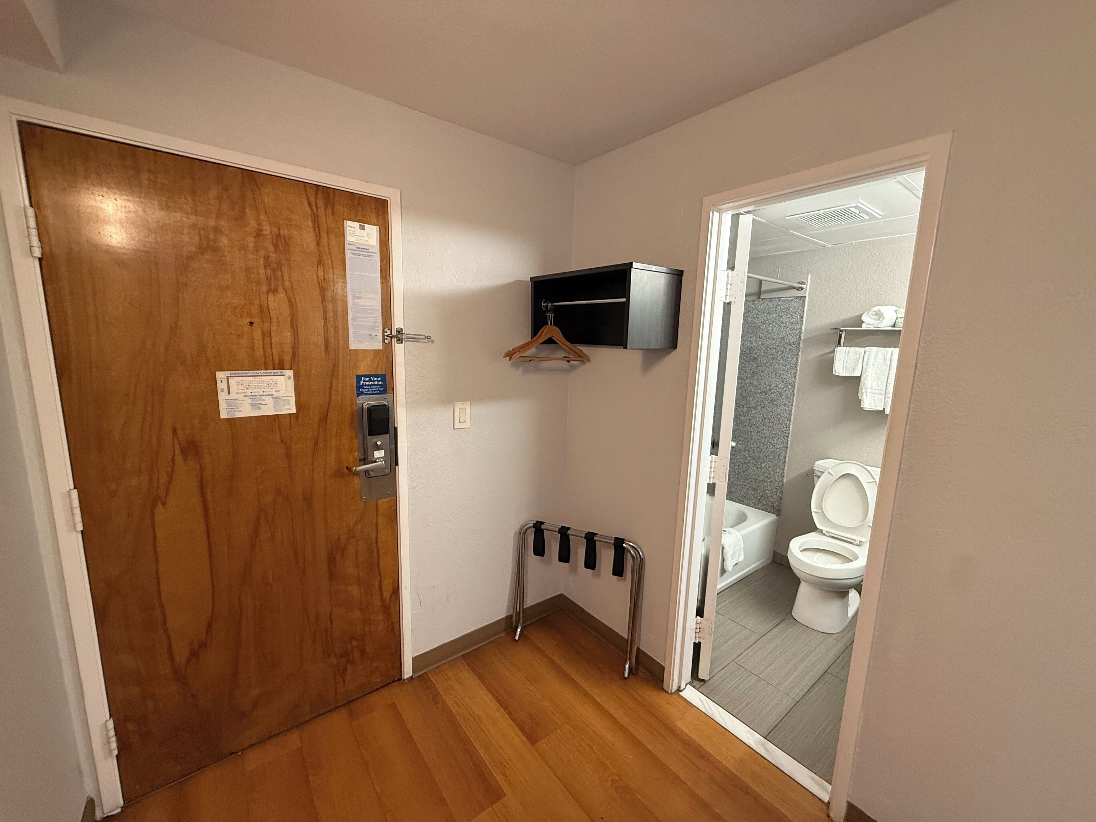 A narrow hallway with a wooden door, a small shelf with a television, and a view into a bathroom with a toilet and a shower.