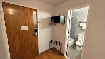 A narrow hallway with a wooden door, a small shelf with a television, and a view into a bathroom with a toilet and a shower.