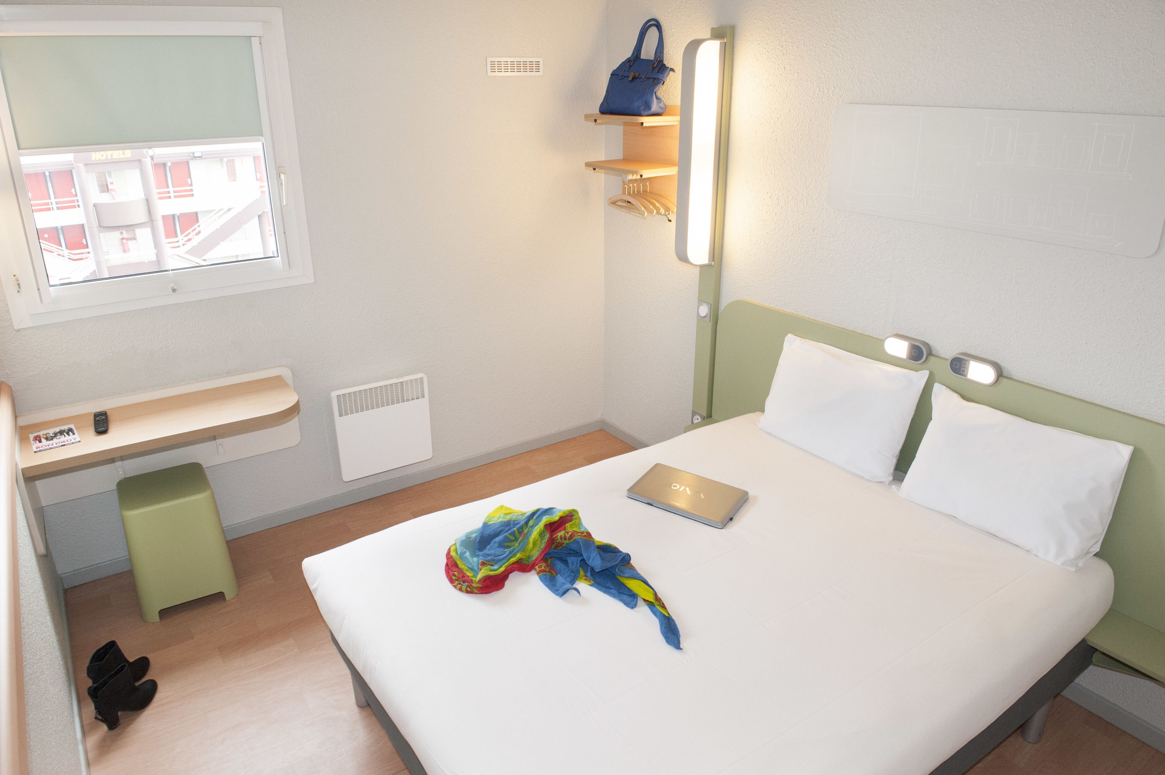 Une chambre d’hôtel avec un lit, un bureau avec un ordinateur portable, une fenêtre et une étagère fixée au mur sur laquelle se trouve un sac à main.