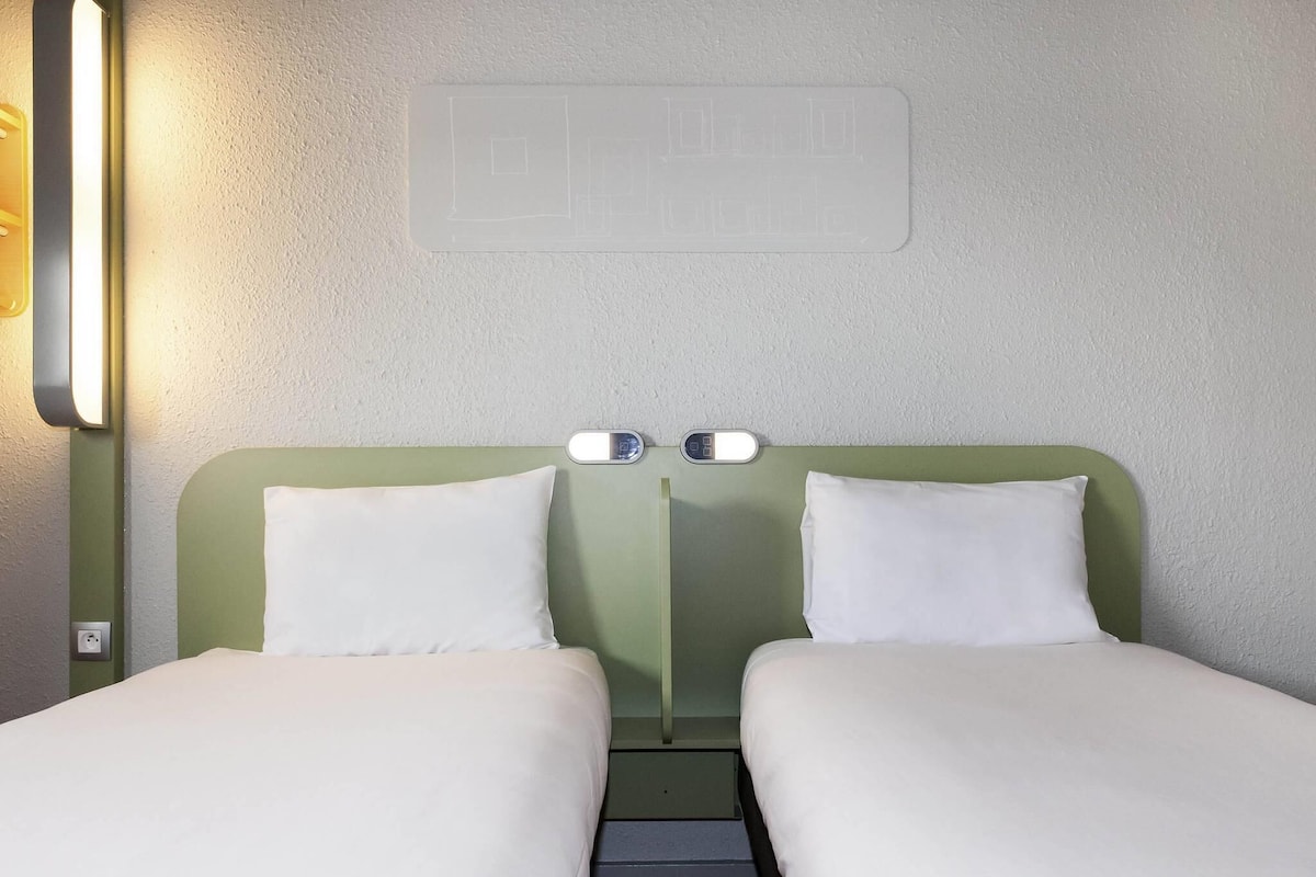 Une chambre double avec deux lits simples, une table de chevet, une lampe fixée au mur et un thermostat.