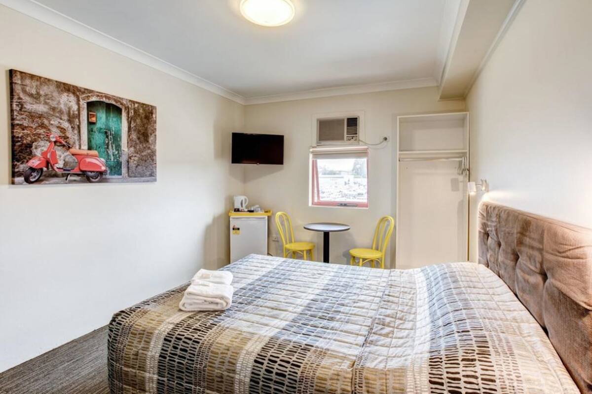 A bedroom with a bed, a painting, a TV, a small table, two chairs, and a window.