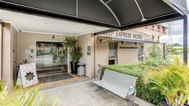 La entrada del motel Sleep Express, con un área de espera cubierta, un banco y un cartel que indica trasbordos de autobús hacia Olympic Park.