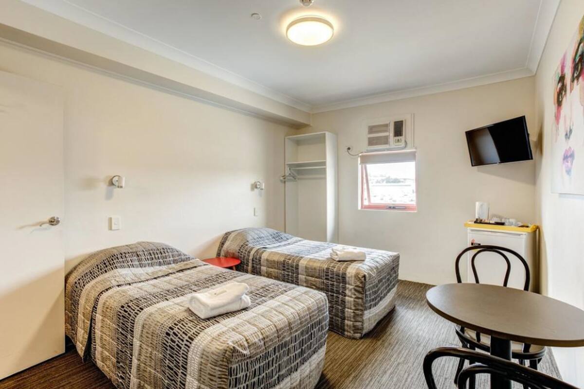 A room with two beds, a small table, a chair, a TV mounted on the wall, and a window with a view.