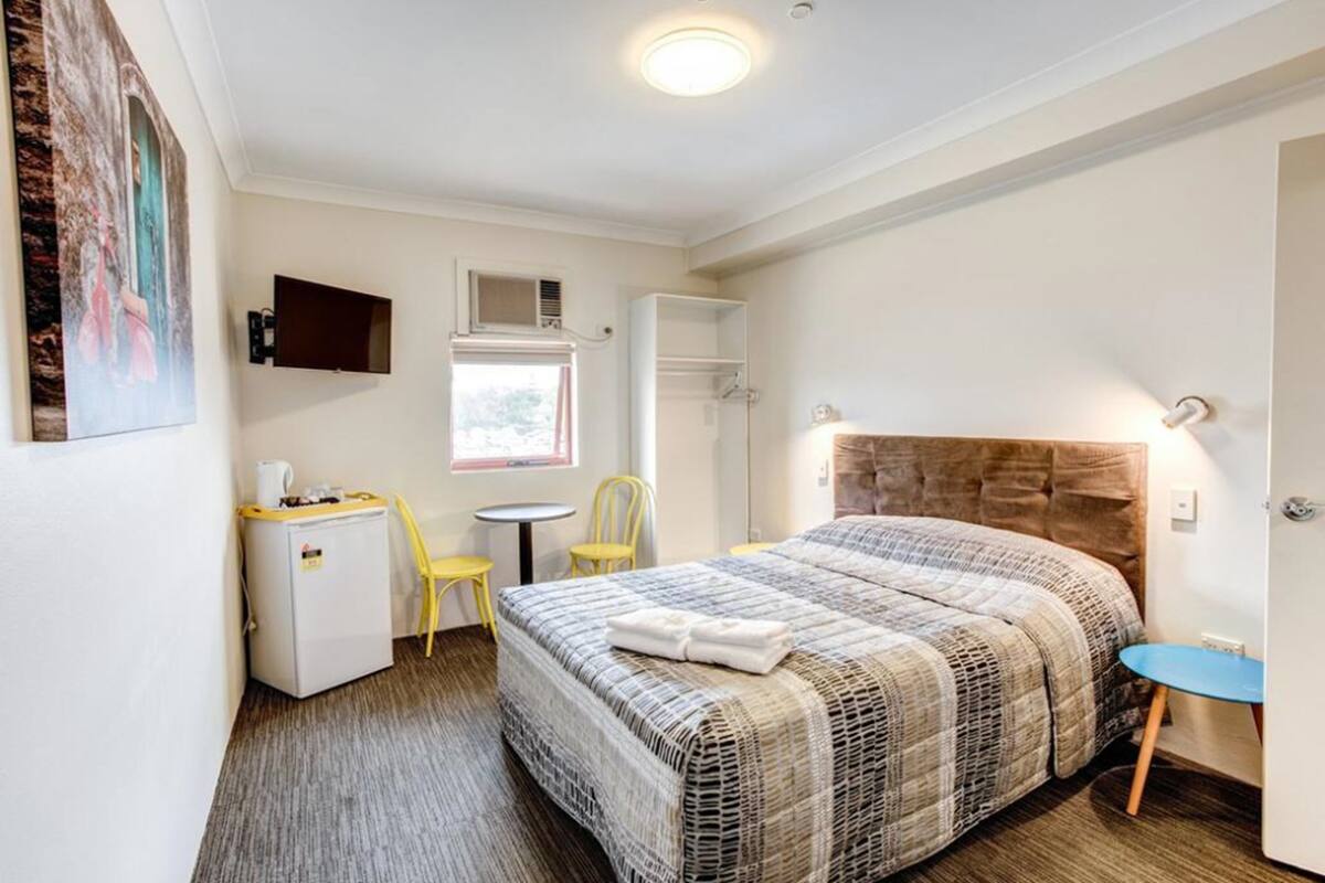 A hotel room with a large bed, a desk with a chair, a small table, and a wall-mounted TV.