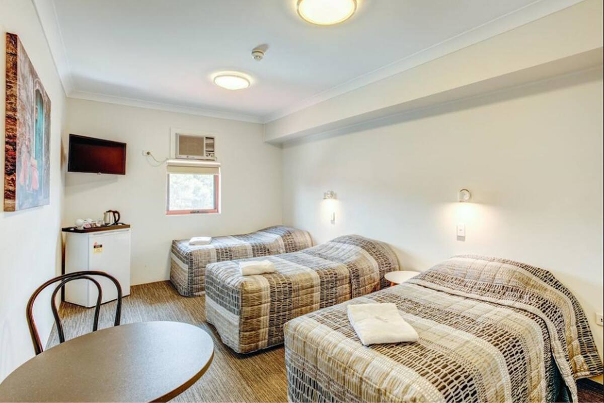 A small, well-lit room with two single beds, a desk with a chair, and a small table.