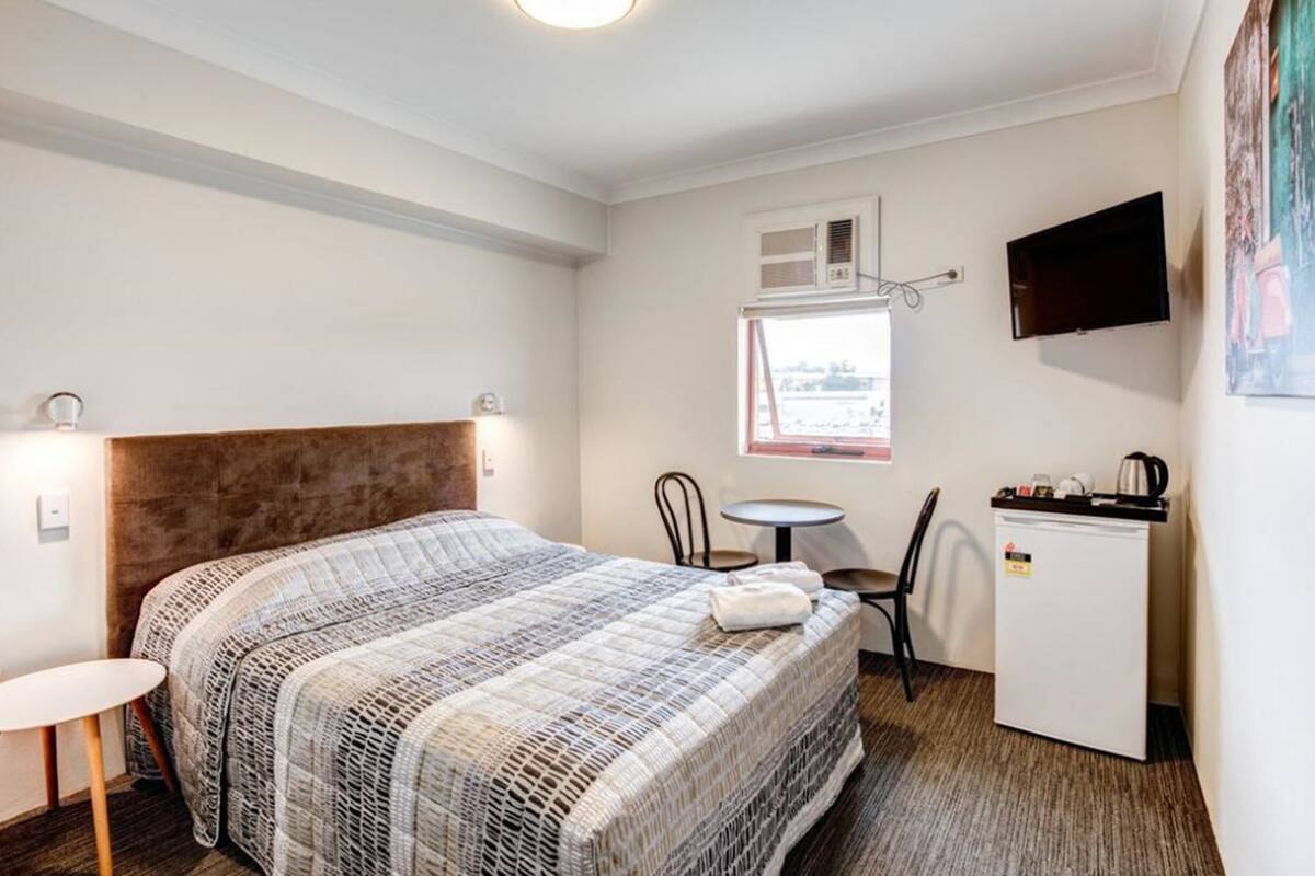 A hotel room with a bed, a small desk, a chair, a television, and a window with an air conditioning unit.