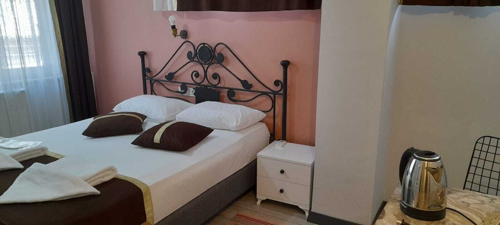 A bed with a metal headboard, white bedding, and two brown pillows. There is a white nightstand with a lamp and a kettle on a table beside the bed.
