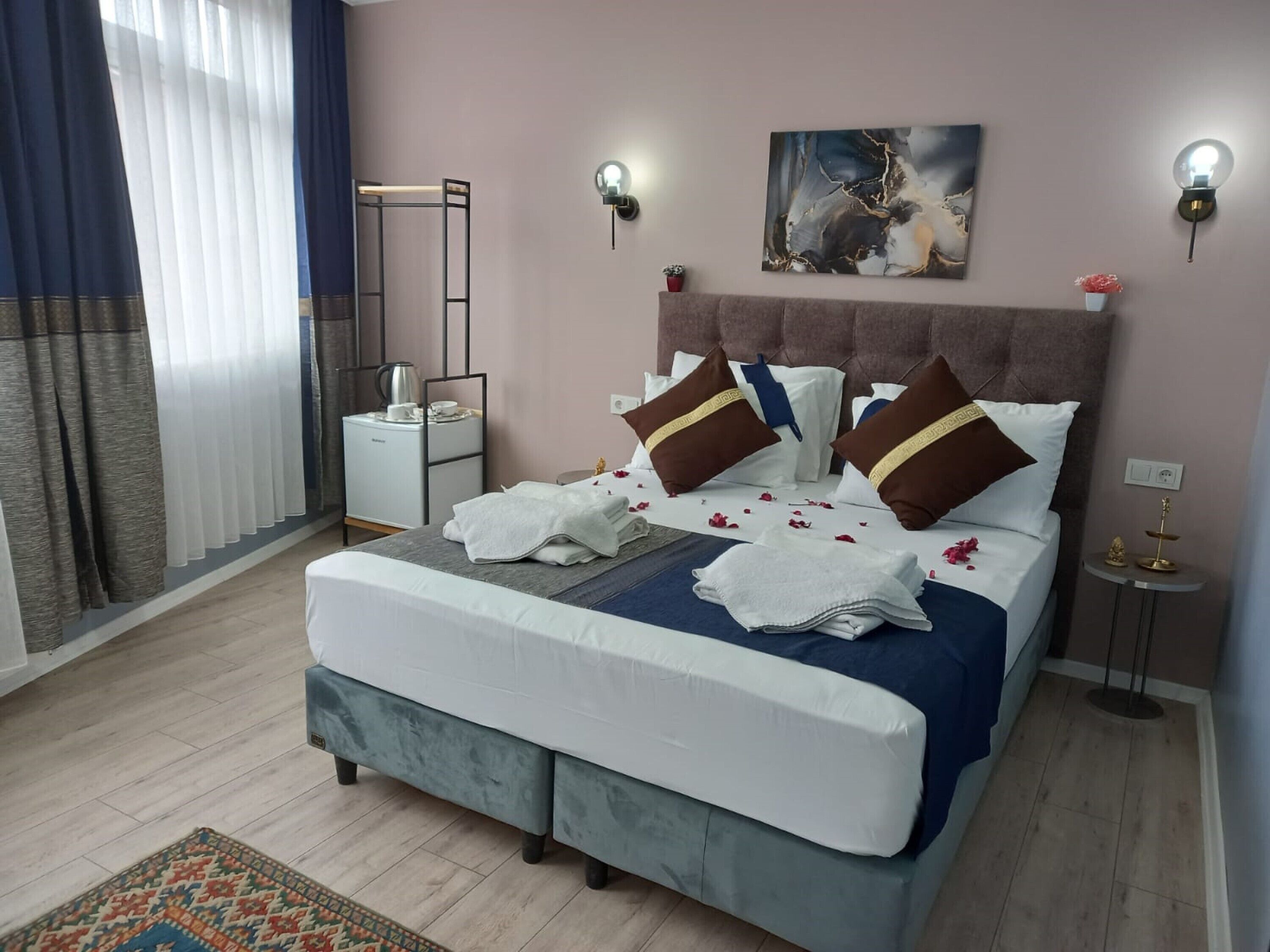 Een net opgemaakt bed met kussens, een nachtkastje met een lamp, een aan de muur bevestigd kunstwerk en een bijzettafeltje met een decoratief object.