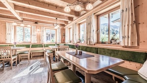 A wooden interior with a long table and benches, large windows with curtains, and a view of greenery outside. - Ferienhof Oberreit (Bruck an der Grossglocknerstrasse)