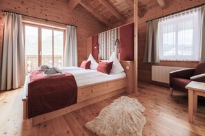 A wooden cabin bedroom with a bed, a chair, a table, and a view of mountains through large windows. - Ferienhof Oberreit (Bruck an der Grossglocknerstrasse)