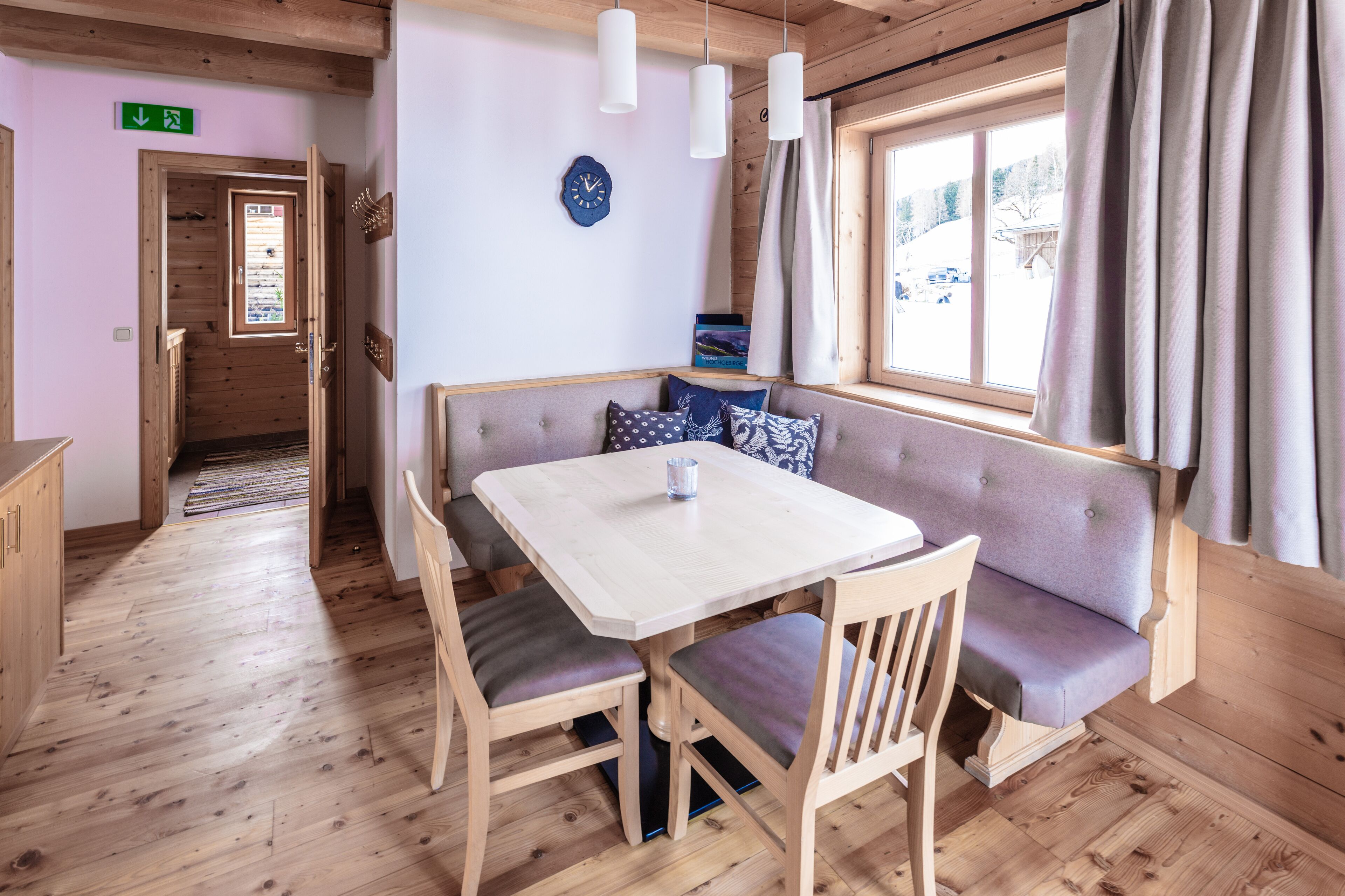 Une pièce au plancher de bois, avec une table à manger, des chaises et un banc. On y trouve une fenêtre avec des rideaux, une horloge au mur et une porte avec un panneau indiquant la sortie.