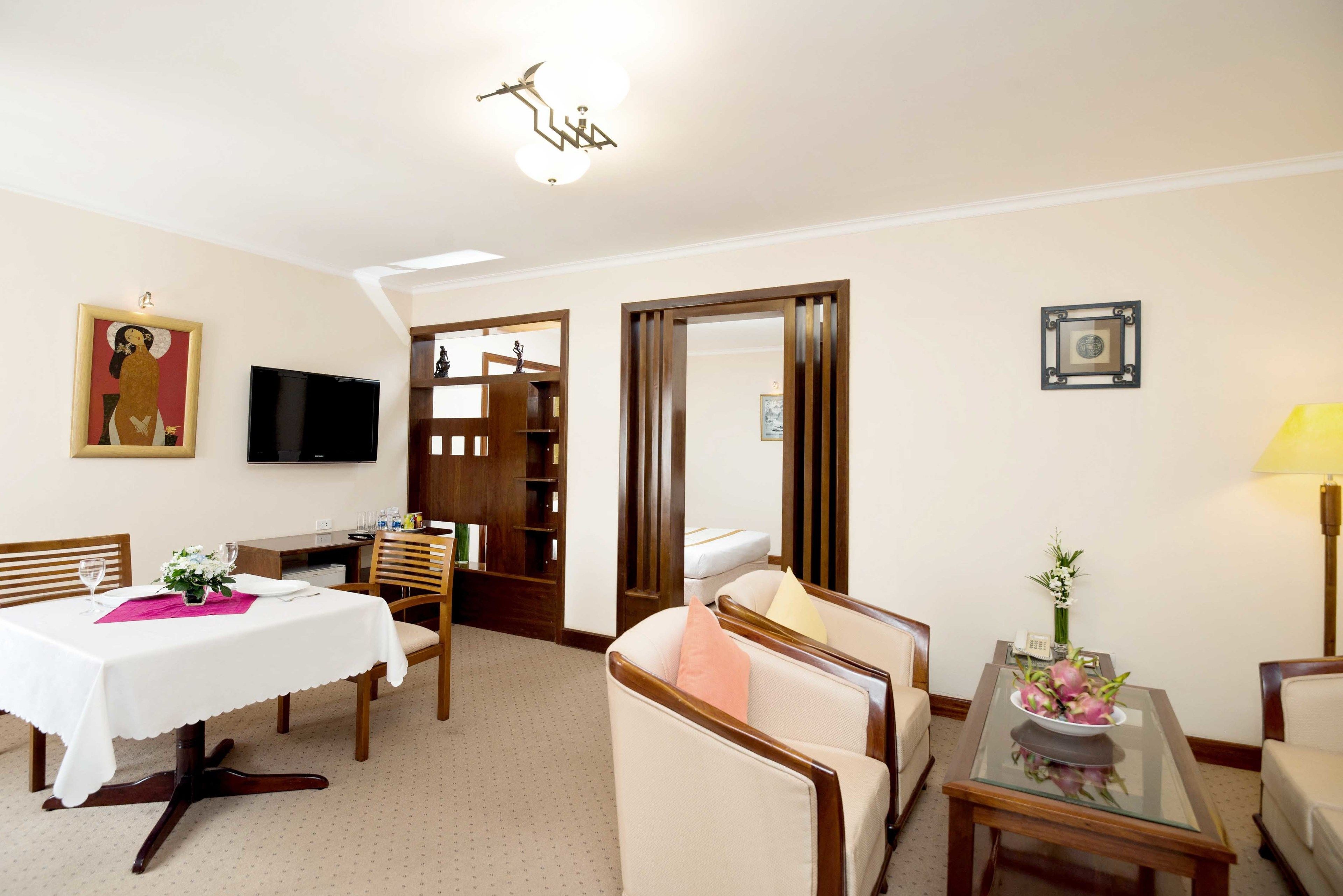 A hotel room with a dining area, a TV, a wooden cabinet, a sofa, and a small table with a vase.