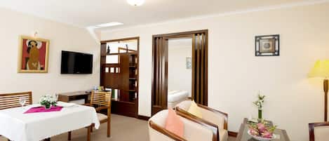 A hotel room with a dining area, a TV, a wooden cabinet, a sofa, and a small table with a vase.