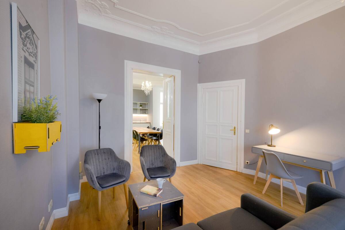 Een moderne woonkamer met een houten vloer, twee grijze fauteuils, een klein tafeltje, een bureau met een lamp, en een witte deur.