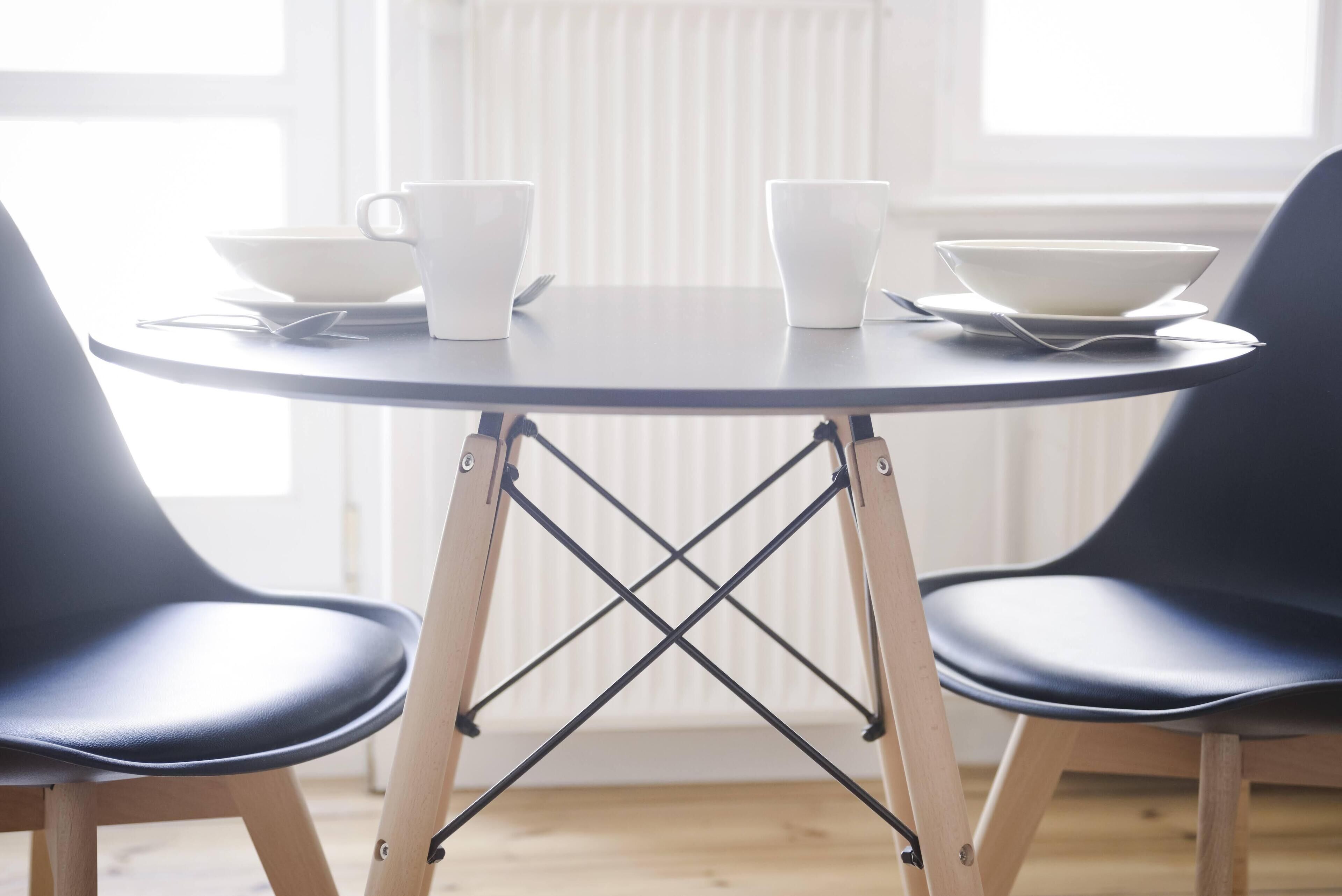 A round table with a white bowl and two white cups on it, flanked by two black chairs.