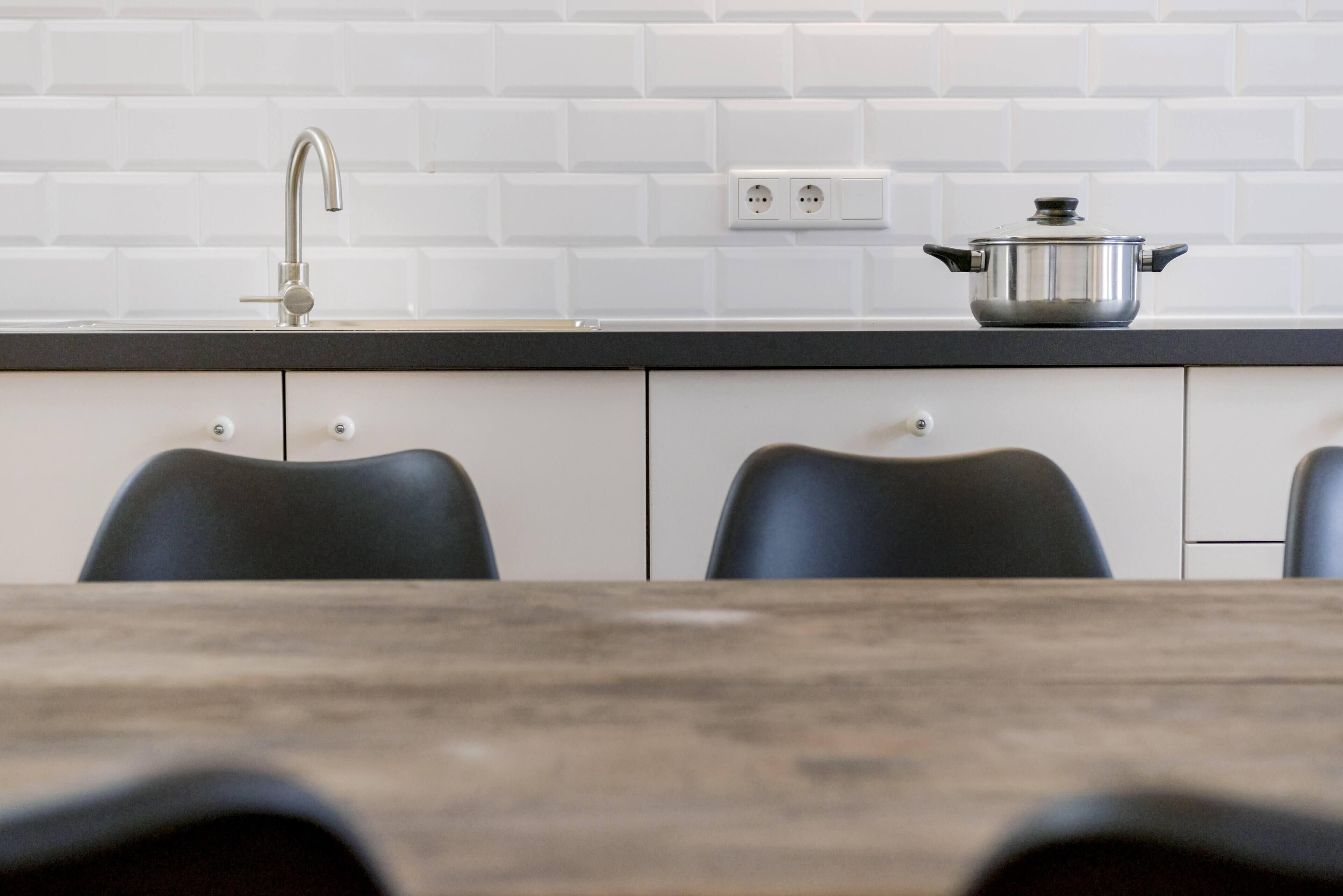A kitchen with white subway tiles, a stainless steel faucet, a pot on the counter, and black chairs around a wooden table.