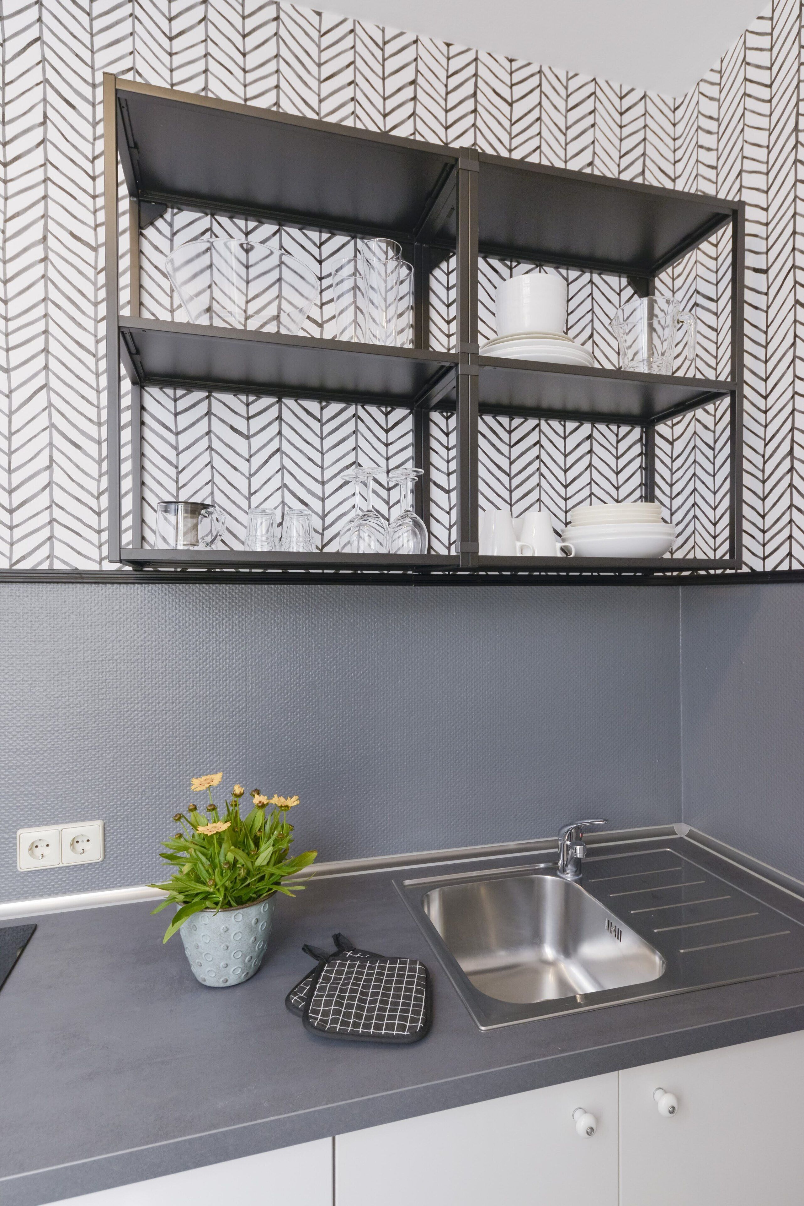 Eine moderne Küche mit grauer Arbeitsplatte, einem Edelstahlspülbecken und einem schwarzen Drahtgestell. Auf der Arbeitsplatte steht ein Blumentopf mit gelben Blüten.