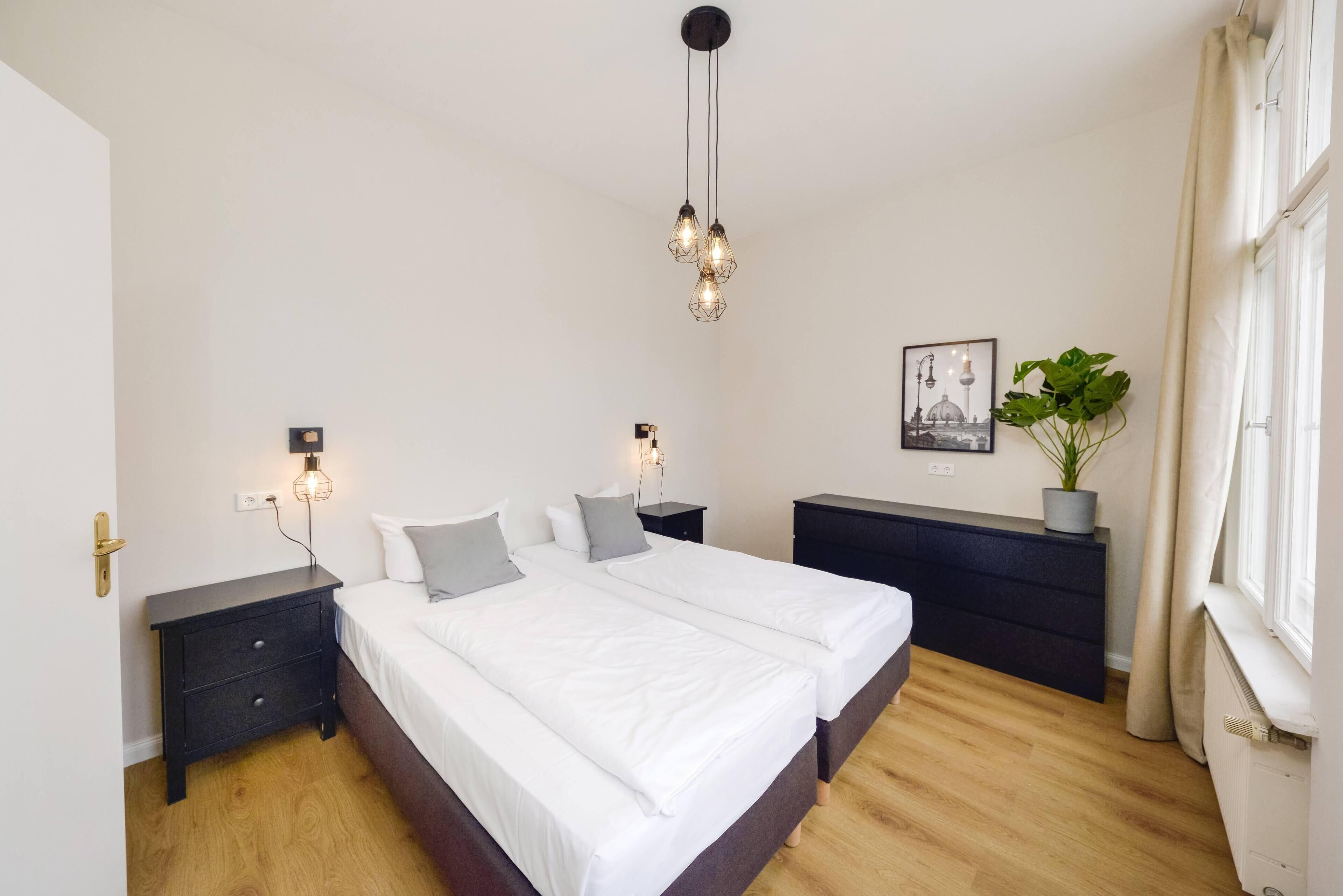 A bedroom with a large bed, two bedside tables, a dresser, a hanging light fixture, a wall-mounted picture, and a potted plant.