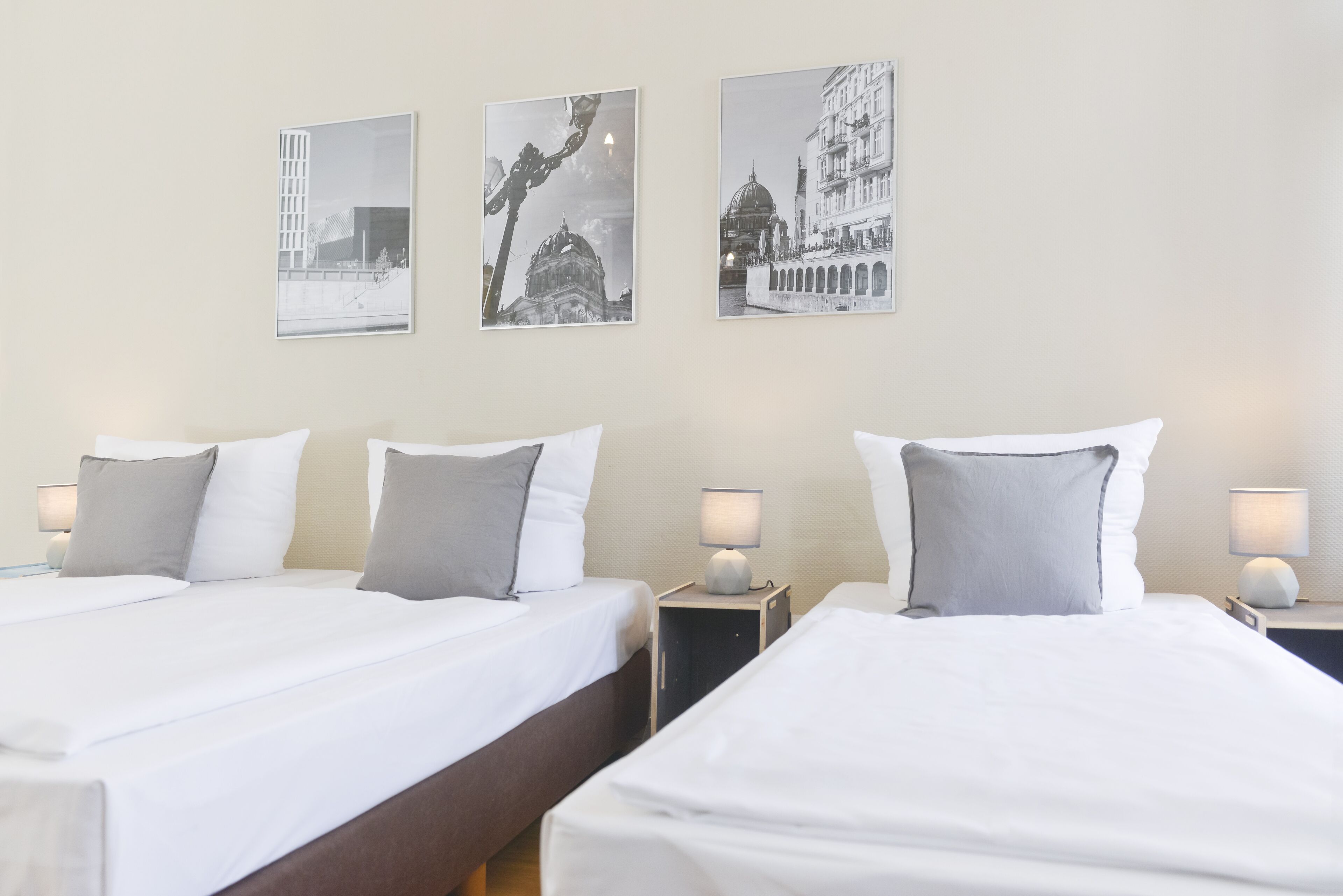 A hotel room with two beds, each with white linens and grey pillows, and bedside tables with lamps. There are three framed pictures on the wall depicting cityscapes.