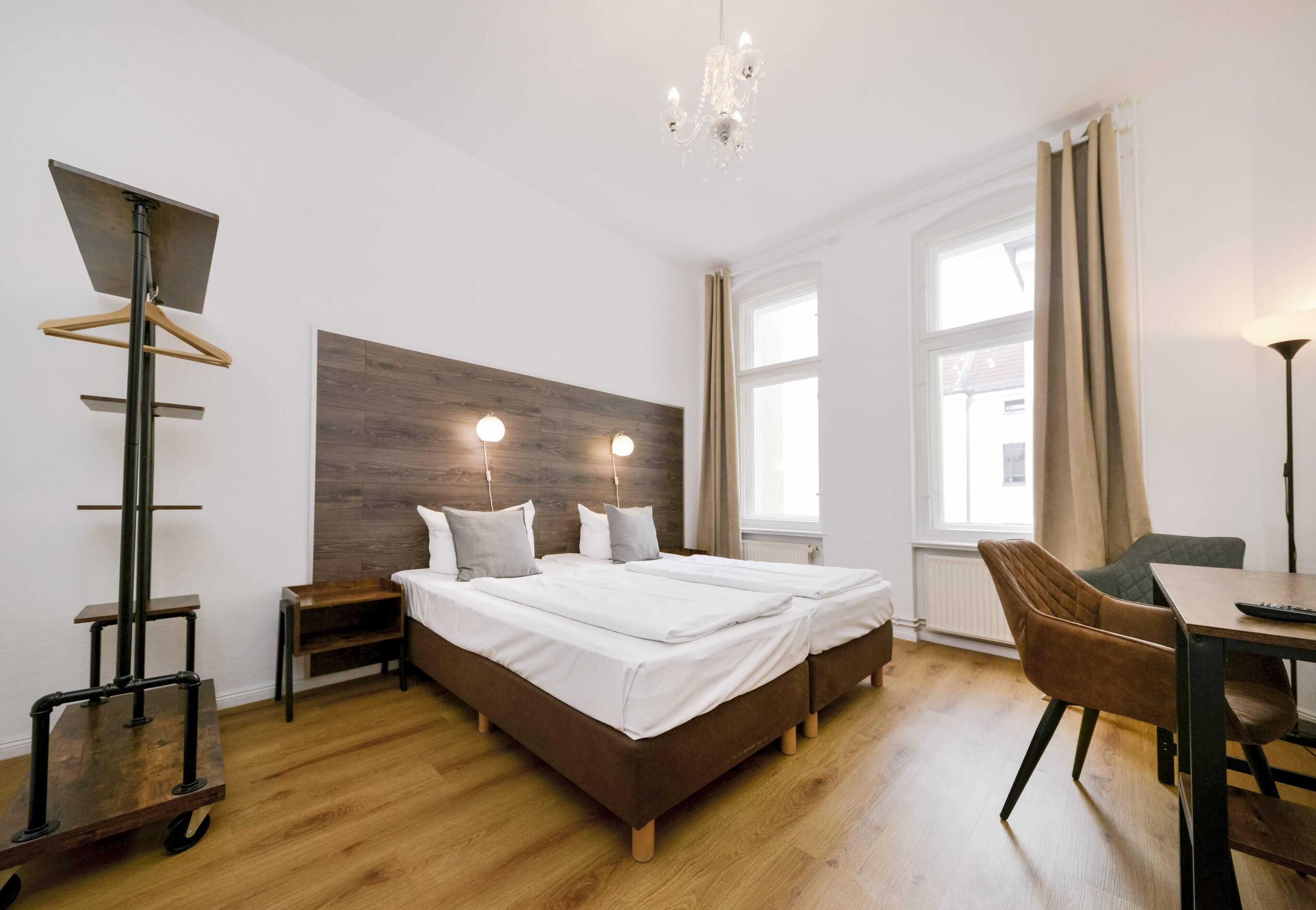 A modern hotel room with a wooden floor, a large bed with white linens, a desk with a chair, and a chandelier.