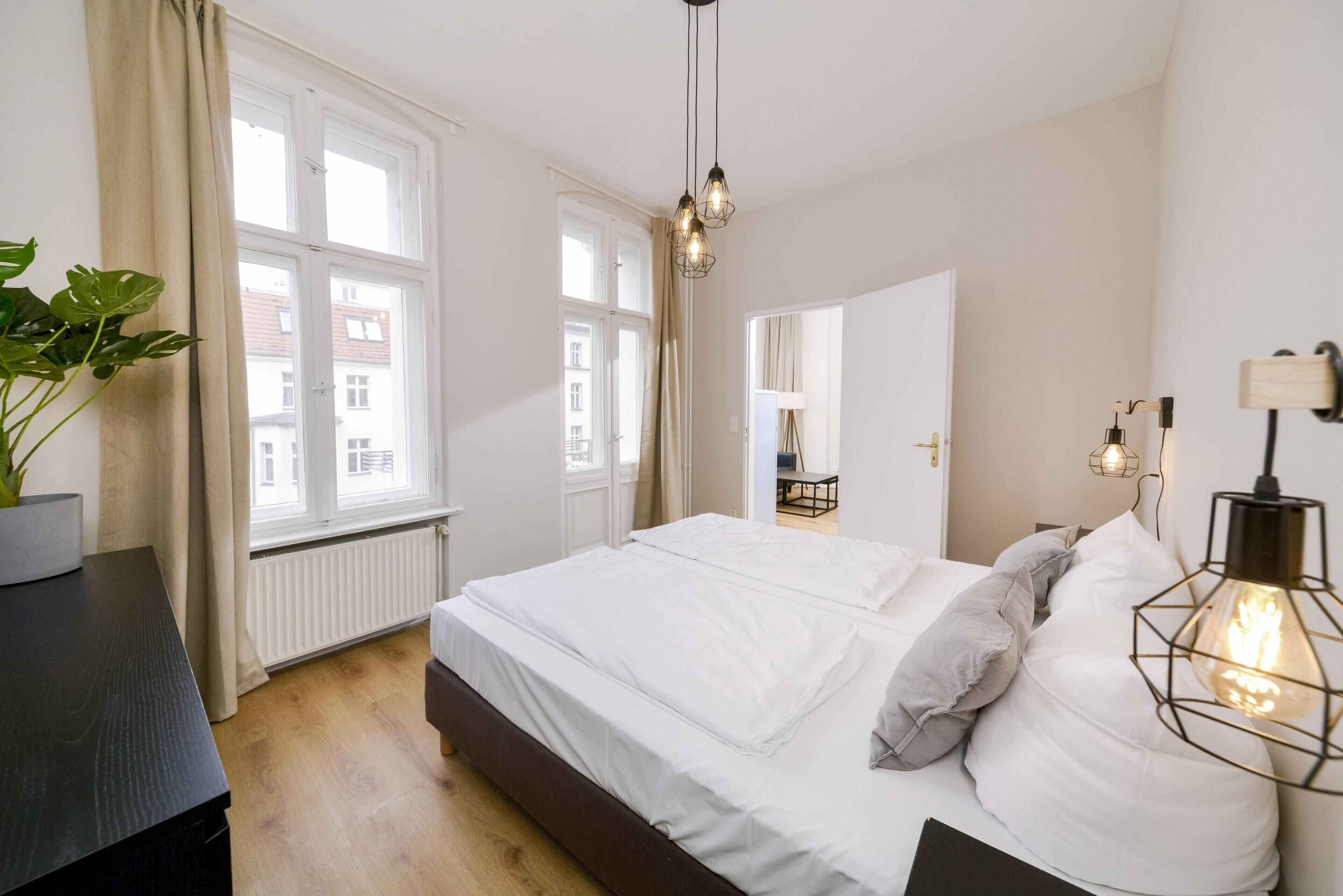 Quarto com uma cama grande, duas luminárias de cabeceira, um vaso com planta e vista para o exterior por uma janela.