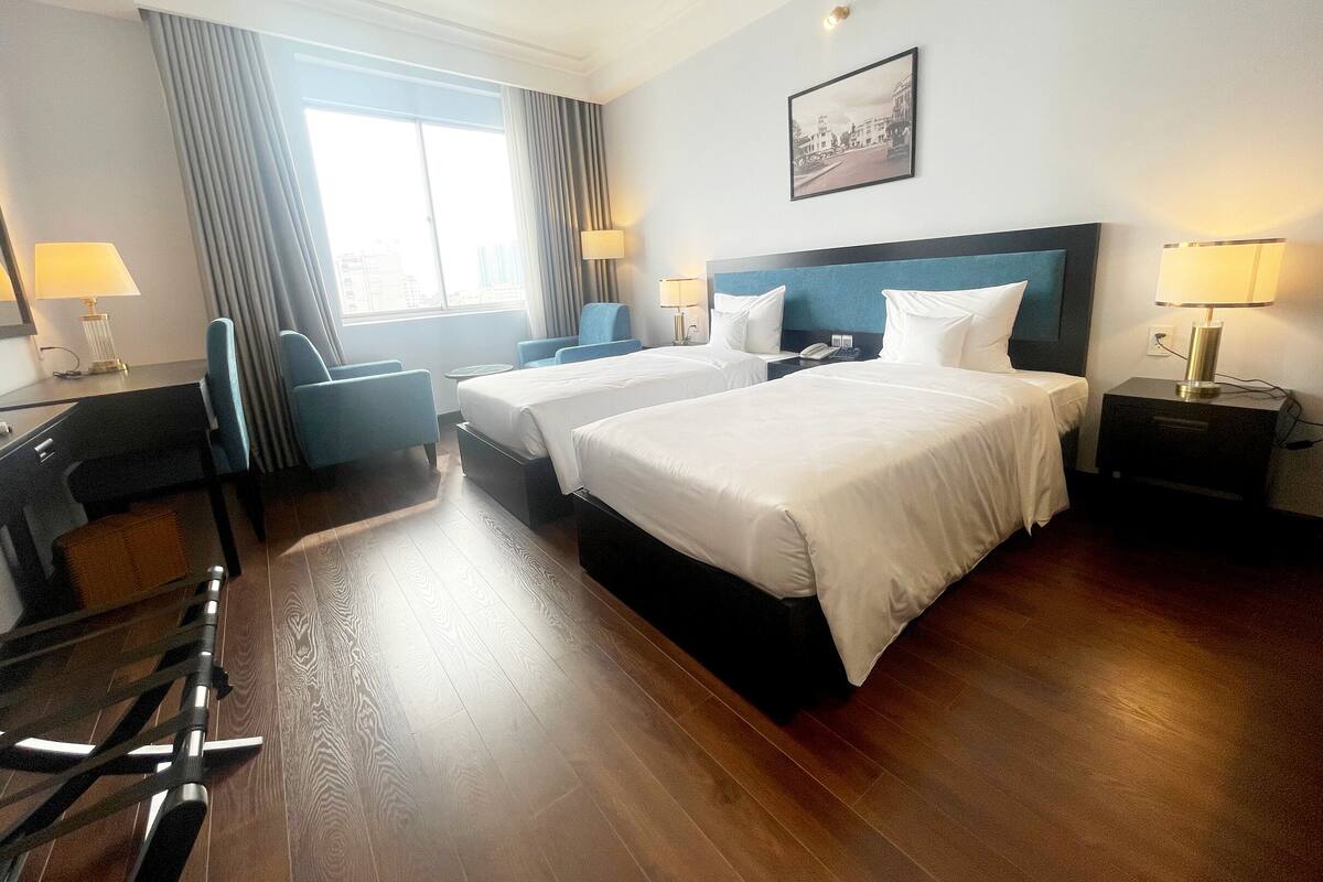 A hotel room with two beds, a desk, a chair, a lamp, and a window with curtains.