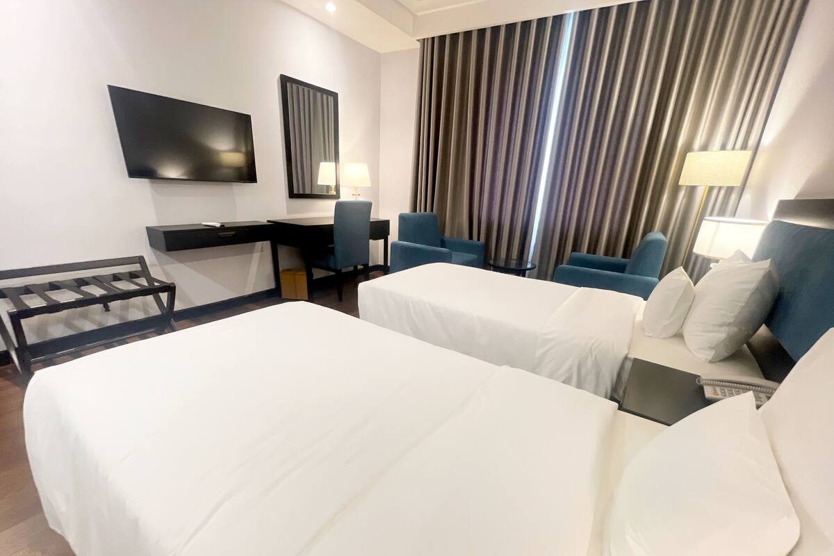 A hotel room with two beds, a desk, a chair, a TV, and a large window with curtains.
