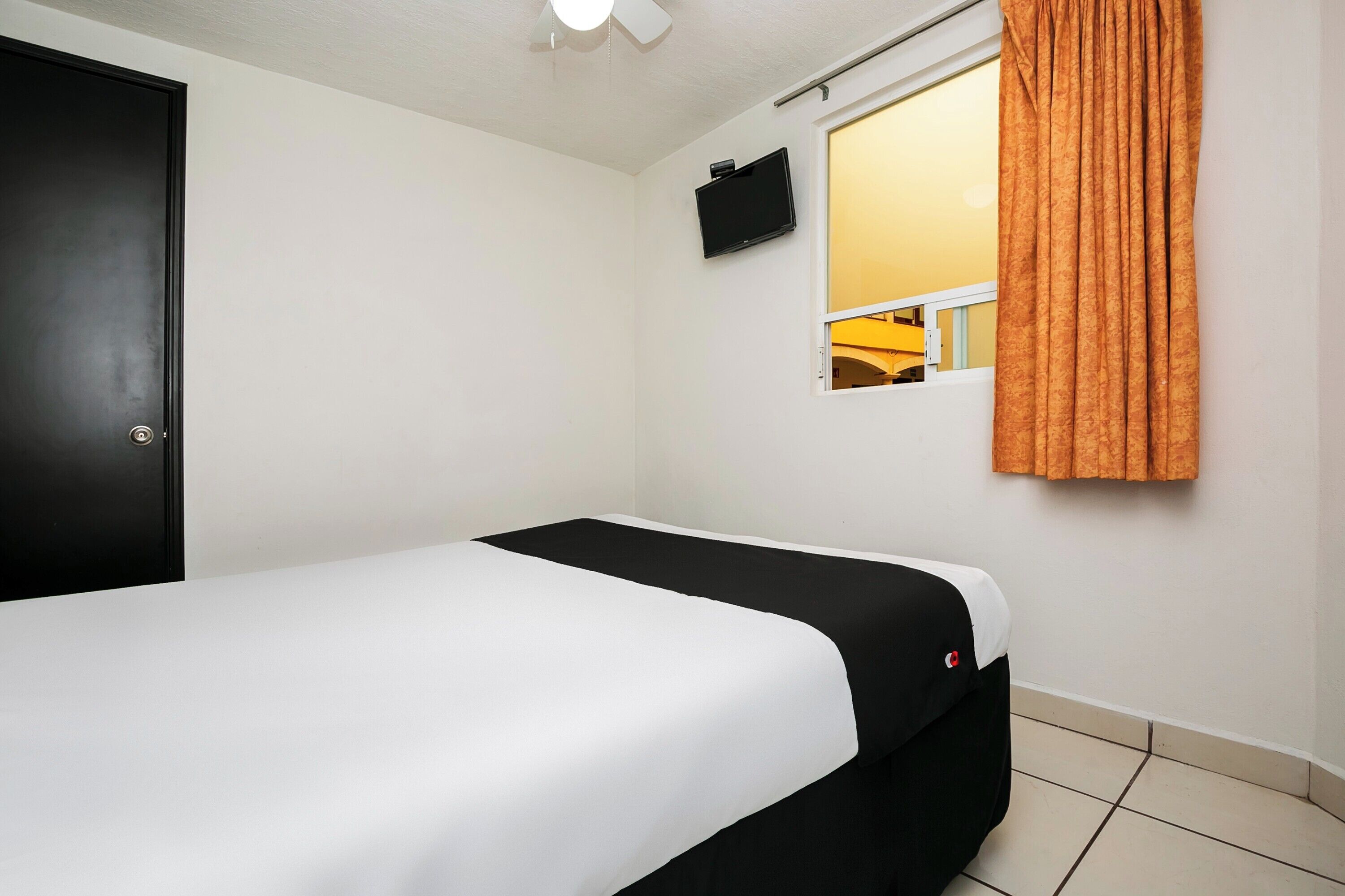 A single bed with a black and white bedspread, a small window with orange curtains, a mounted TV, and a door.