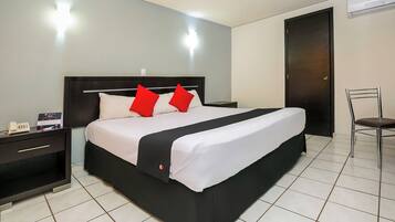 Una cama bien hecha con cabecera negra y ropa de cama blanca, una mesita de noche con un teléfono, y una silla a la derecha.