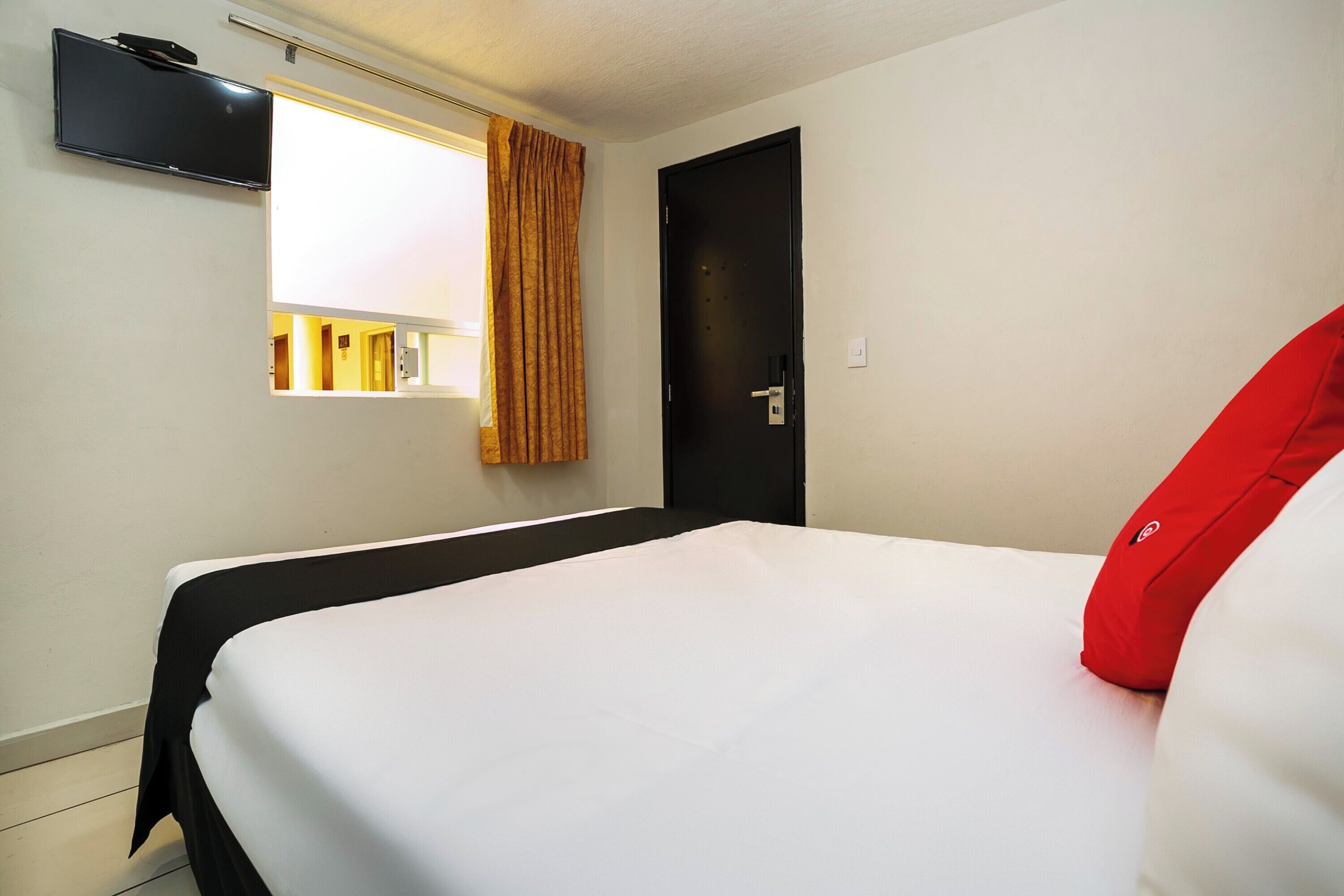Une chambre d’hôtel avec un lit, une fenêtre avec des rideaux, une porte et un téléviseur à écran plat fixé au mur.