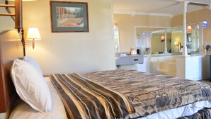 A hotel room with a bed, a nightstand, a lamp, a painting on the wall, a bathroom with a mirror, and a sink. - Knights Inn Greenville, TX (Greenville)