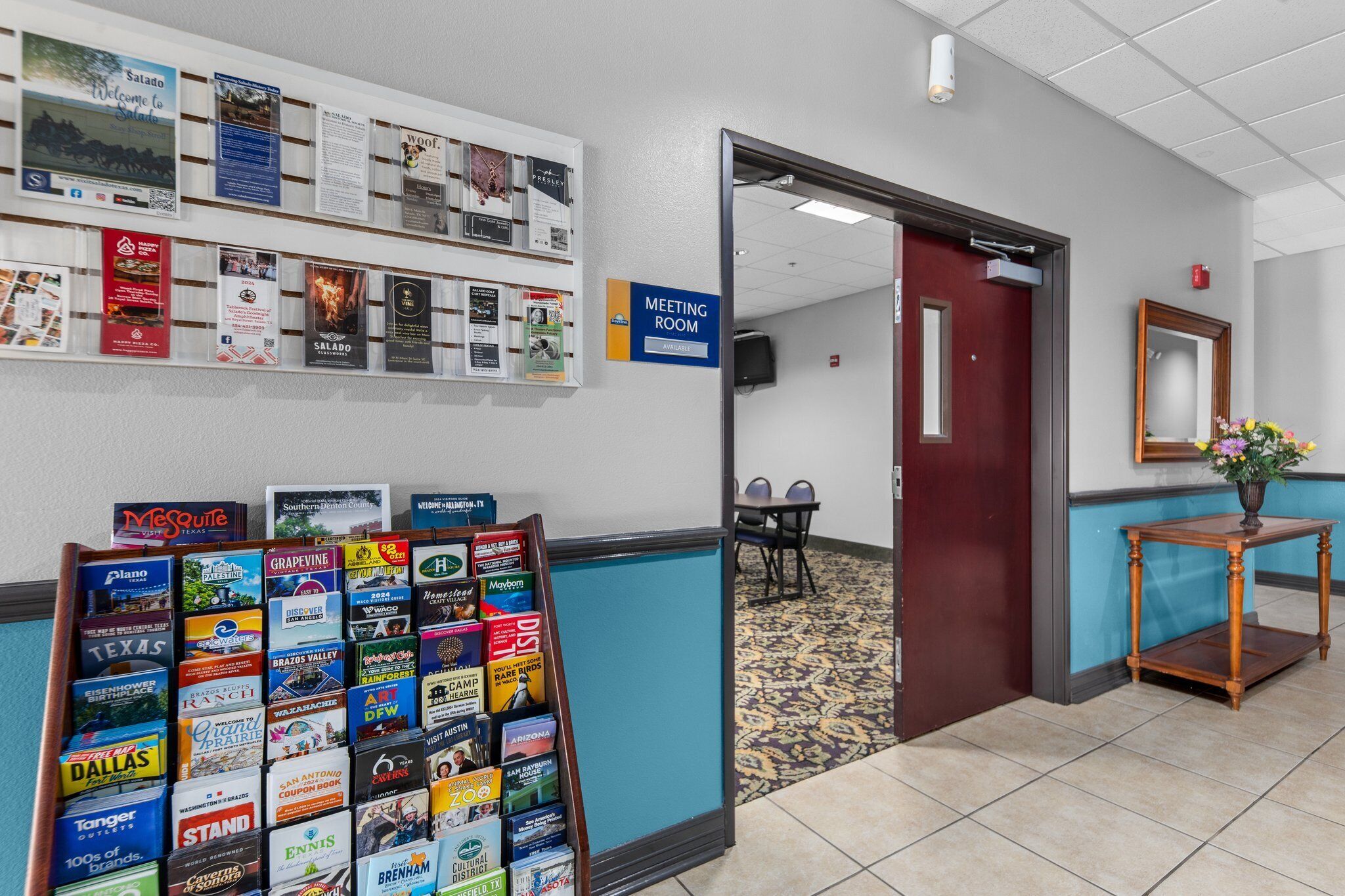 Een hotel lobby met een receptiebalie, een bord met daarop 'Meeting Room' en een muur met diverse brochures en informatieposters.