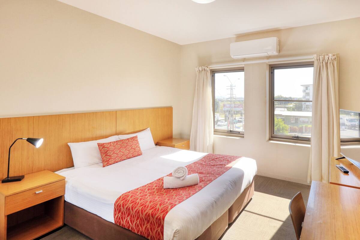 A hotel room with a bed, bedside table, lamp, TV, and a window with curtains.