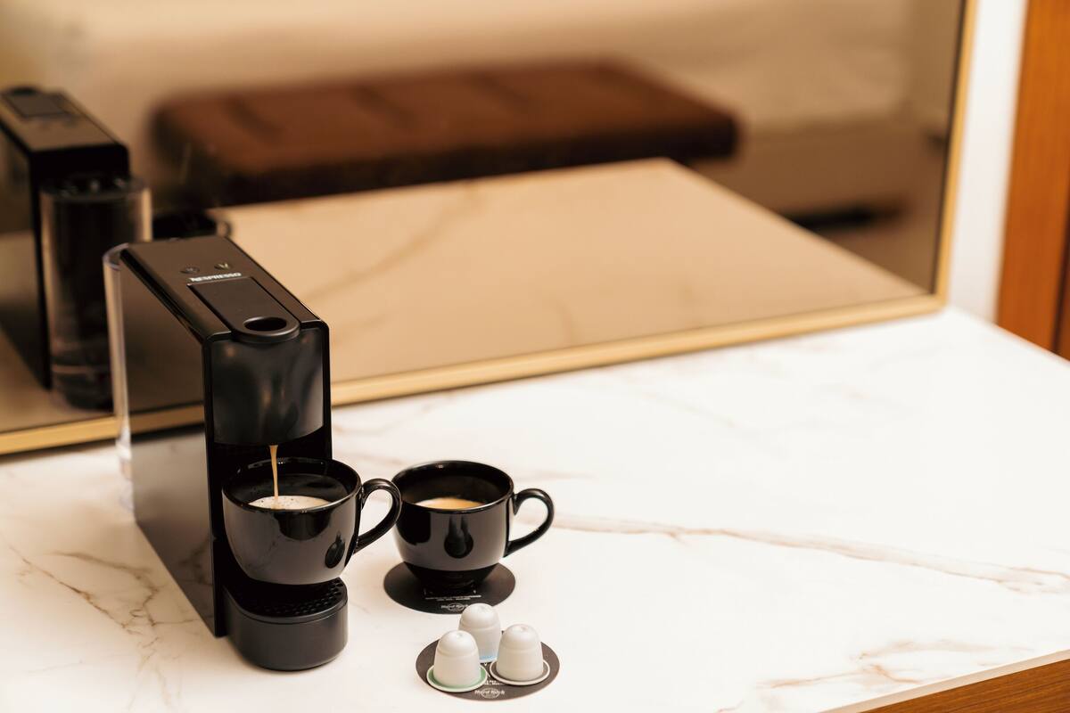 Eine Kaffeemaschine auf einer Marmorarbeitsplatte mit zwei schwarzen Tassen und zwei Kaffeekapseln.