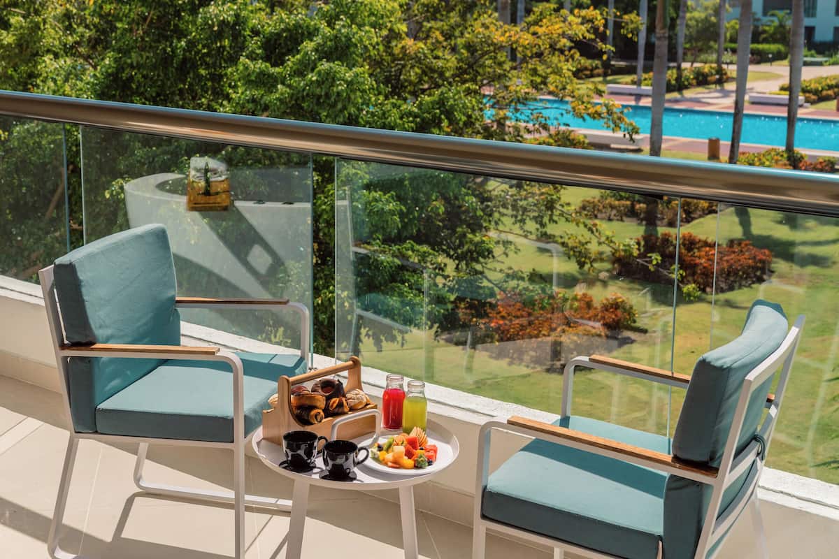 Ein Balkon mit zwei Stühlen, einem kleinen Tisch mit Frühstücksangeboten und Blick auf einen Pool und Grünflächen.