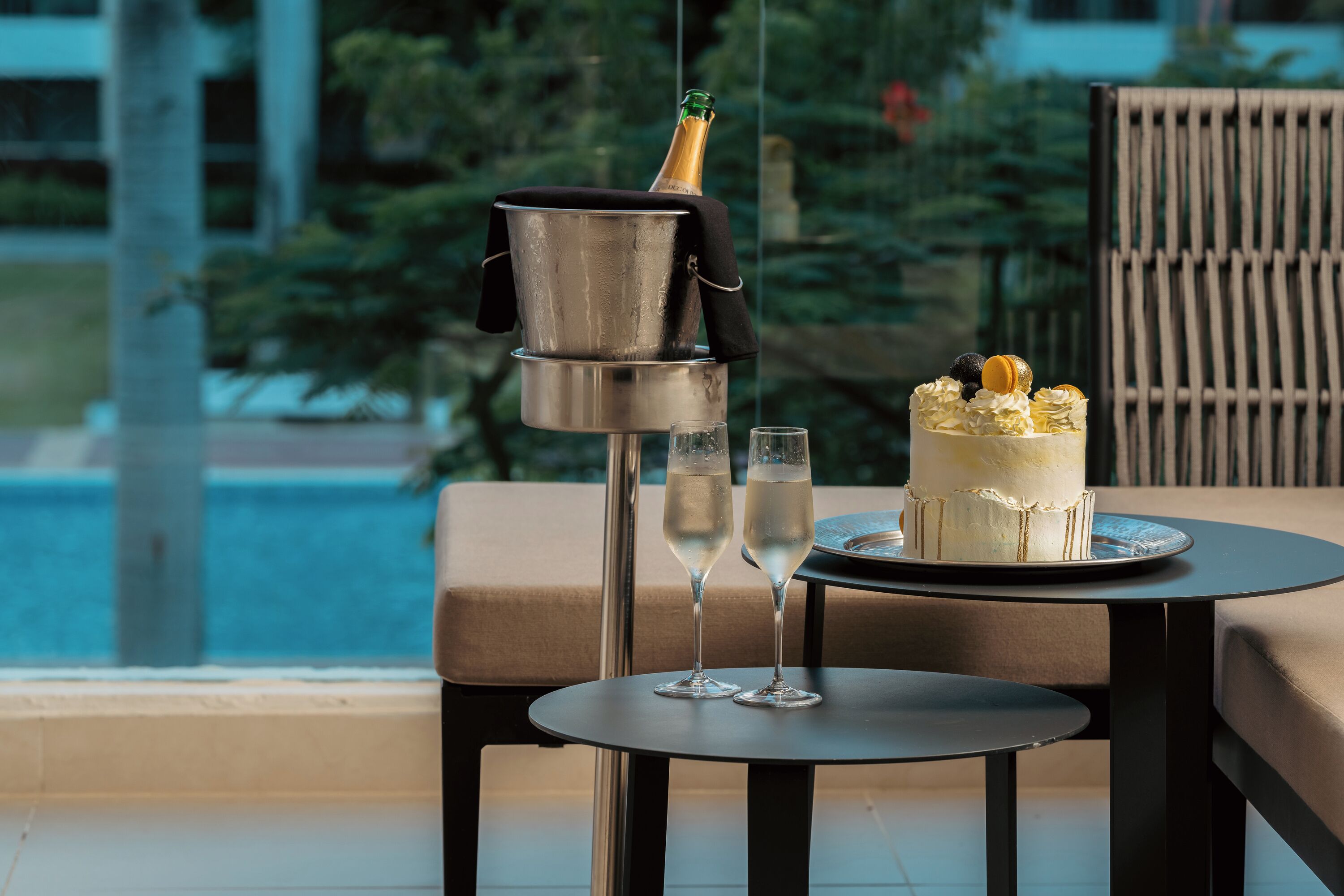 A table with a cake and two glasses of champagne, a metal bucket with a bottle, and a view of a pool and greenery.