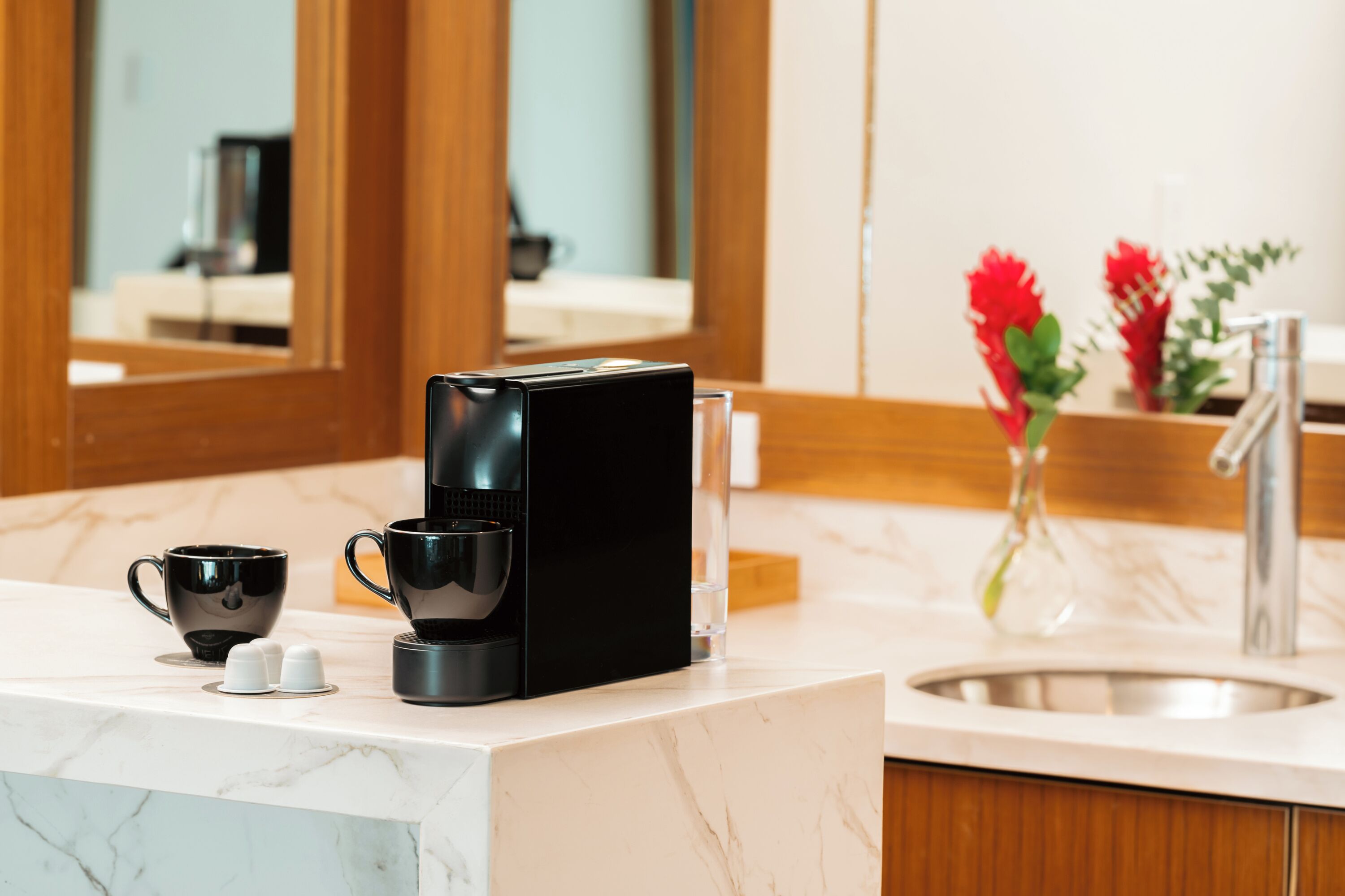 Un bagno moderno con un top in marmo, una macchina da caffè, due tazze nere e un vaso con fiori rossi.