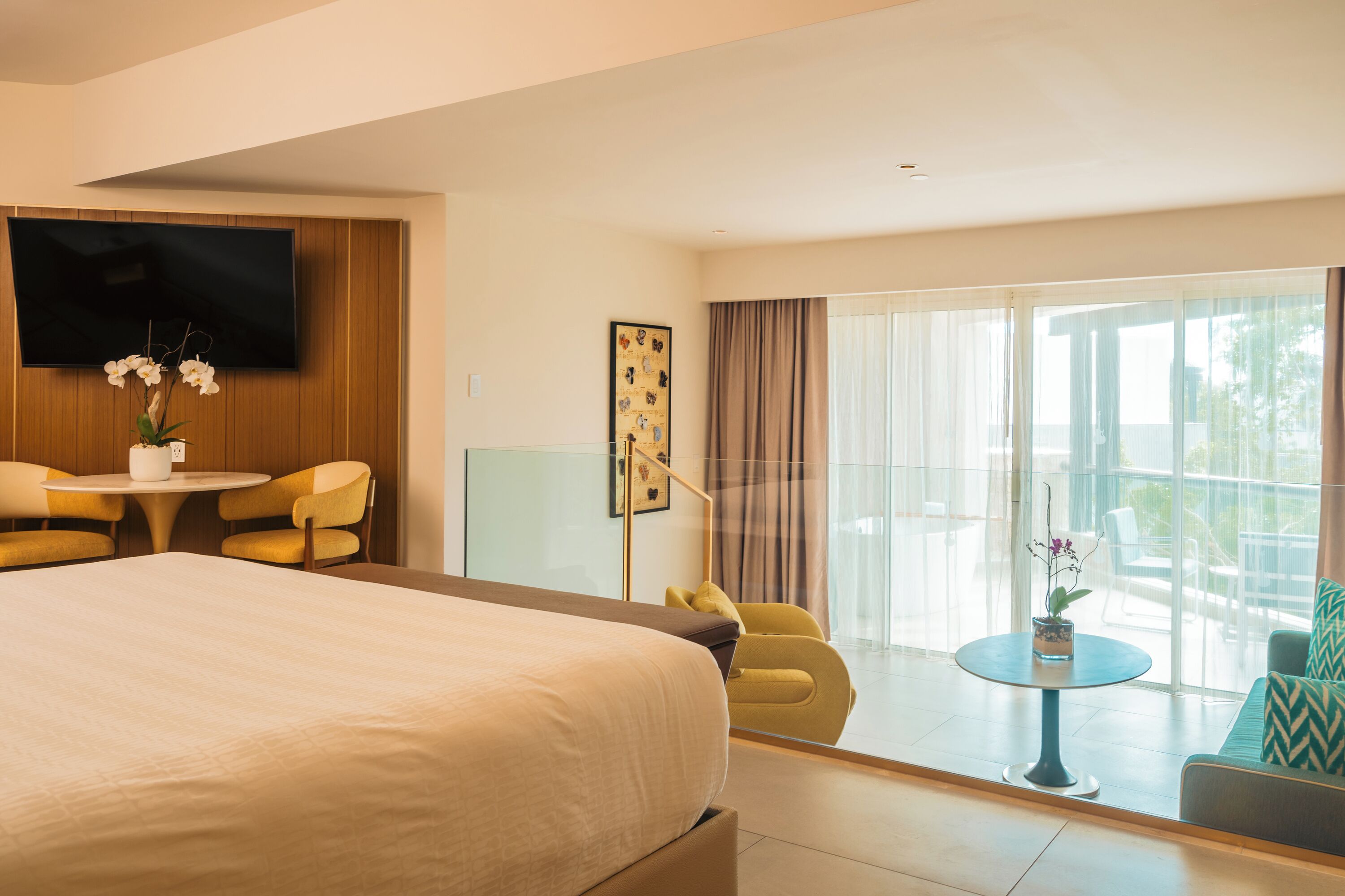 Habitación de hotel con una cama grande, un televisor en la pared, una mesa de comedor con sillas y un balcón con vistas.