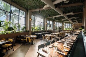 Un restaurante con amplios ventanales, plantas de interior y varias mesas para comer.
