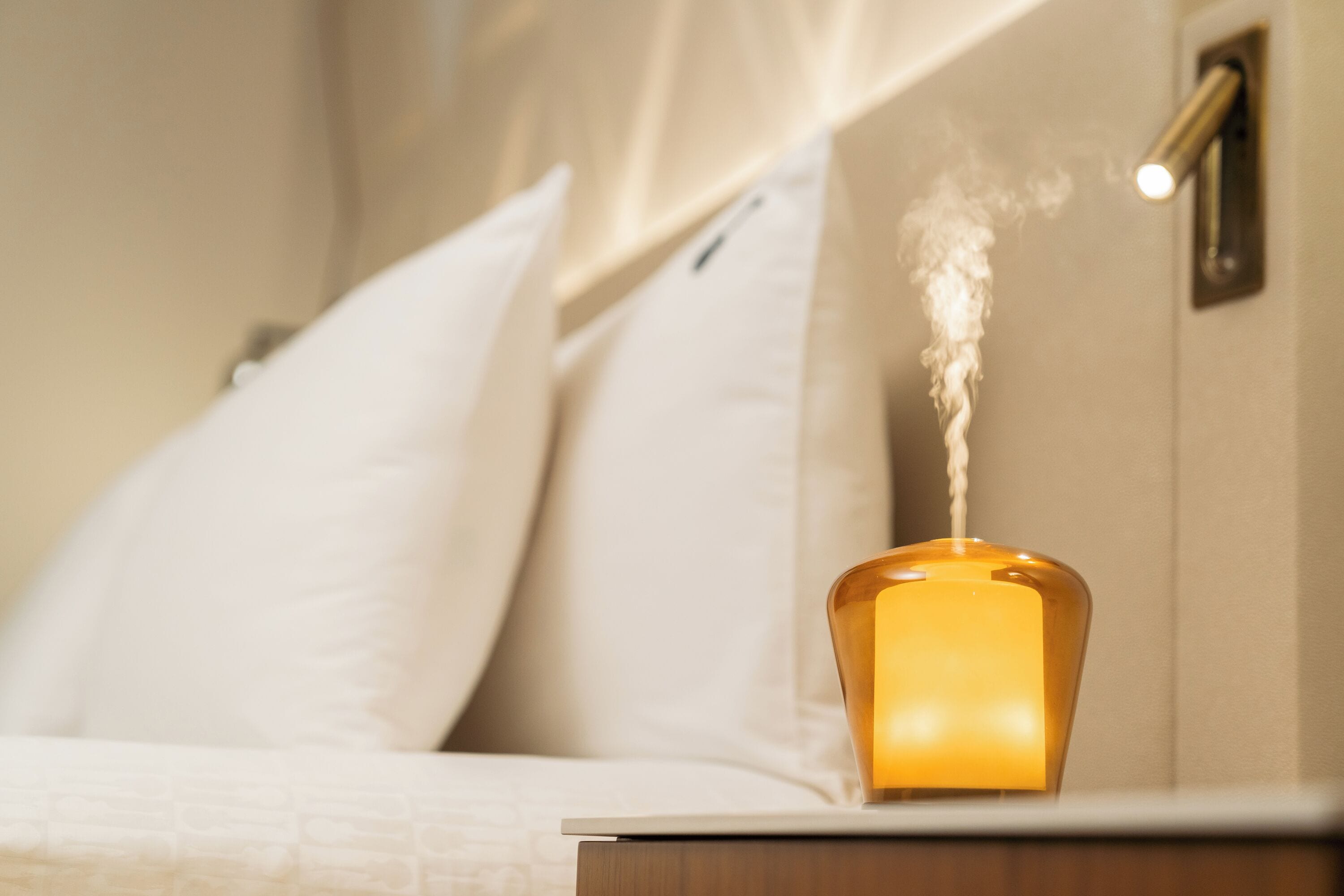 A bedside table with a candle emitting smoke, a white bed with a textured blanket, and a wall-mounted light fixture.