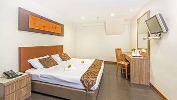 Quarto de hotel com cama, mesa de cabeceira, cadeira, escrivaninha e televisão fixada na parede.